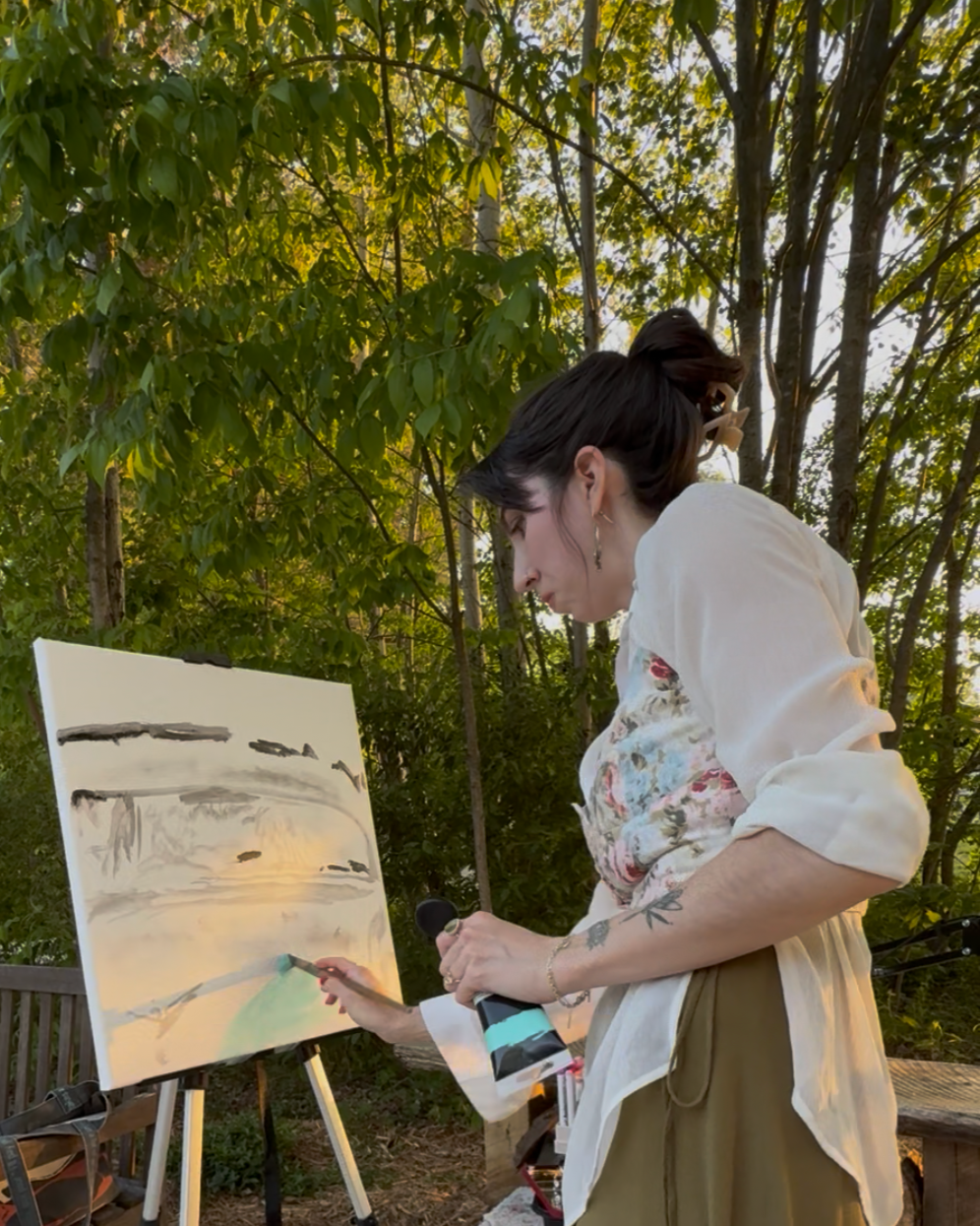 A woman painting outdoors at sunset or sunrise, surrounded by trees.