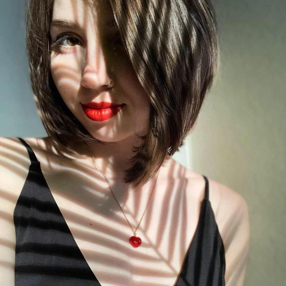 A woman with short brown hair, red lipstick, and a silver nose ring, wearing a black spaghetti strap top and a red heart pendant necklace, standing in front of a wall with light and shadow patterns of a palm leaf