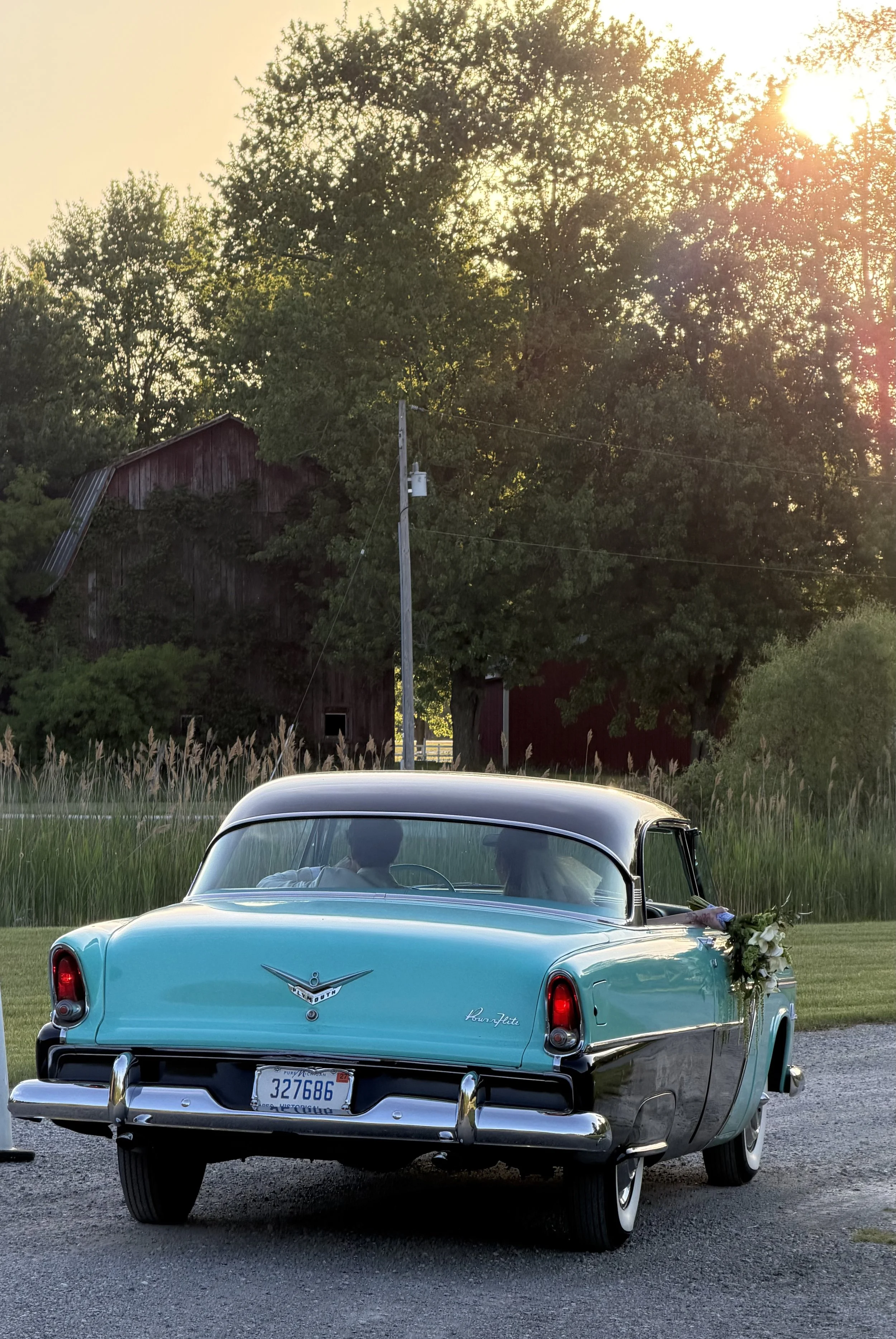 A vintage turquoise Plymouth automobile with a white roof, parked on a grassy area near a pond, decorated with flowers on the rear bumper, with trees, a barn, and a sunset in the background.
