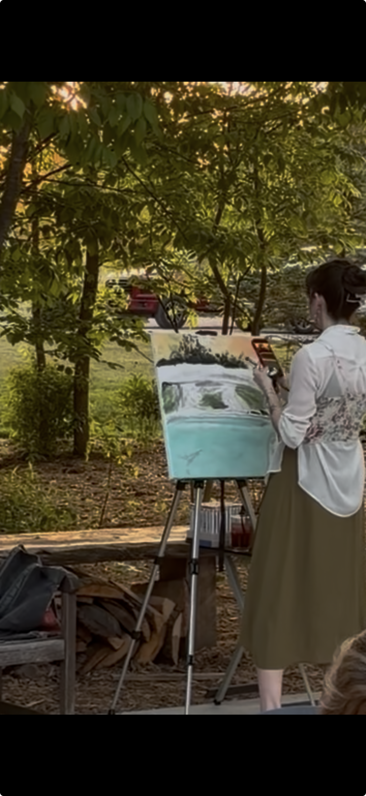 A woman in a white jacket and long skirt is painting outdoors on an easel, surrounded by trees and greenery, with a hose and parked vehicles visible in the background.