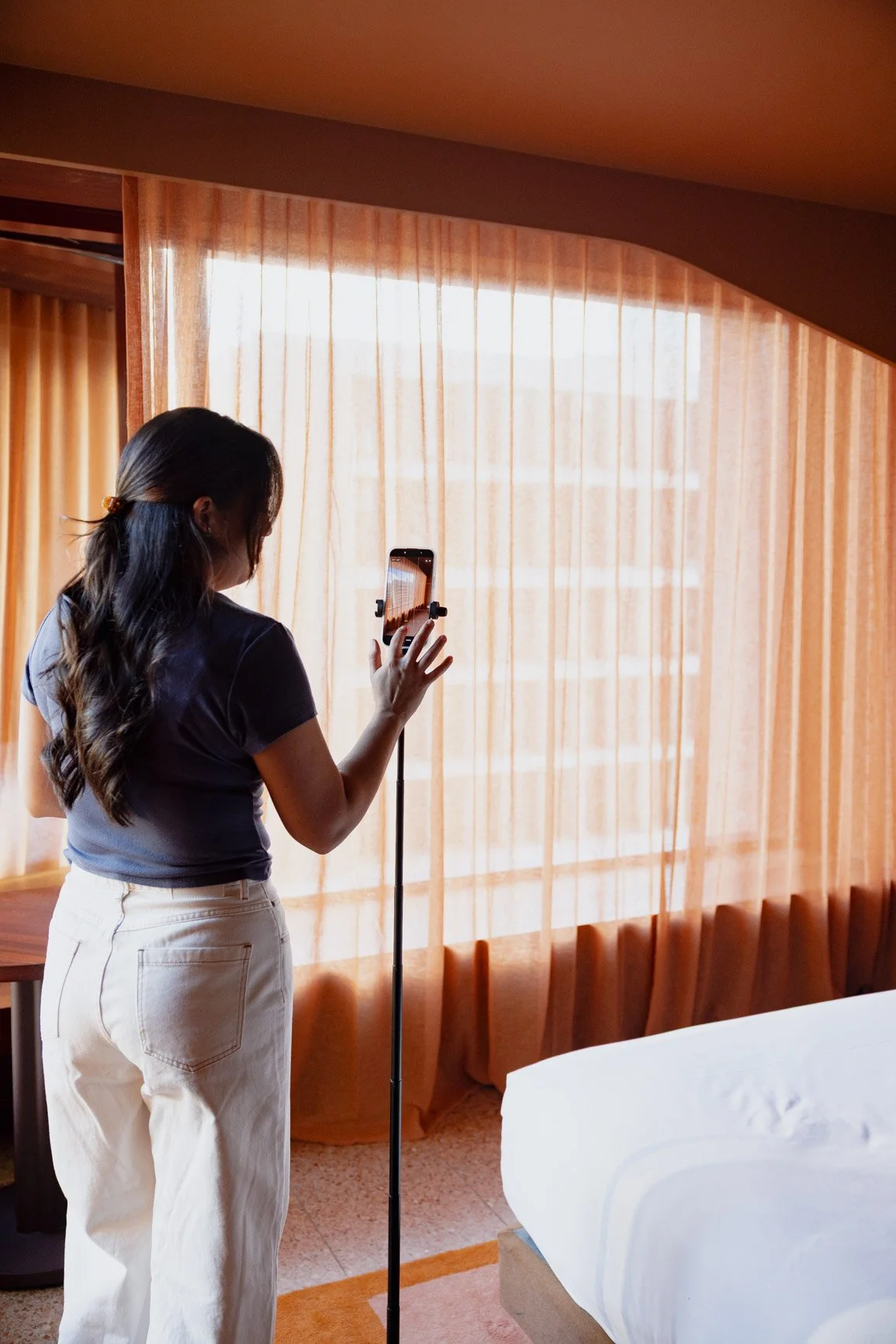 A woman standing in a hotel room, taking a photo with her smartphone.