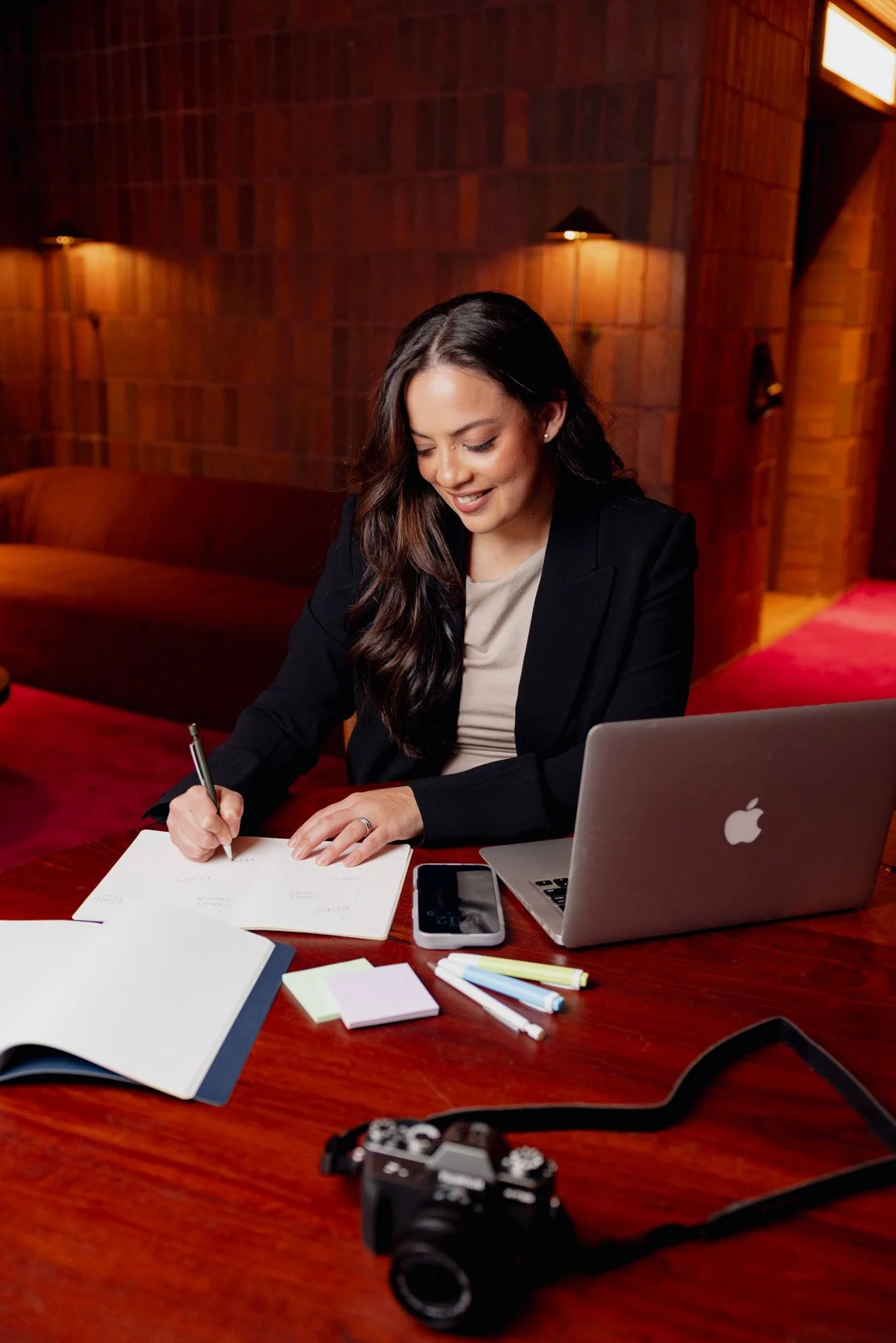 A woman with long, dark hair writing in a notebook at a wooden desk with a laptop, smartphone, sticky notes, and pens, in a warmly lit room with wooden walls.