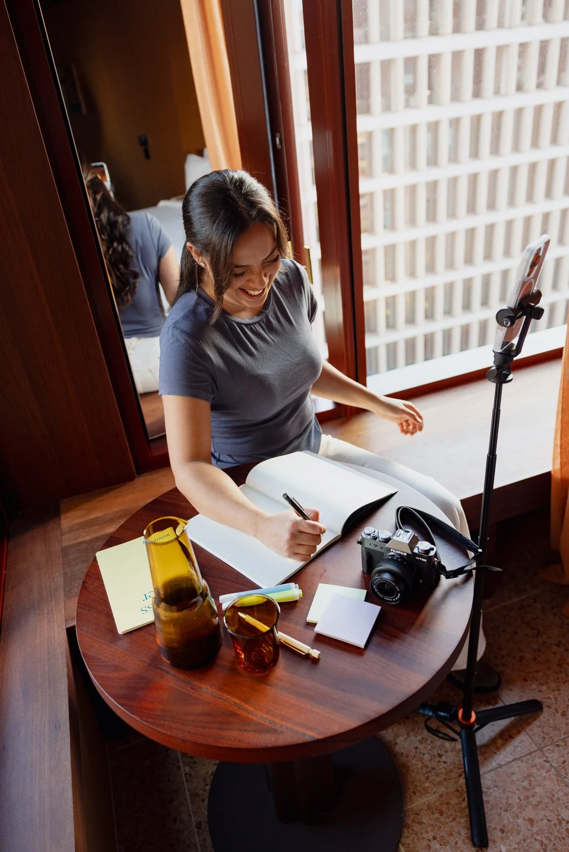 A woman sitting at a round wooden table, smiling and writing in a notebook, with a camera, a glass, a notebook, and a pen on the table, near a large window with a wooden railing outside.