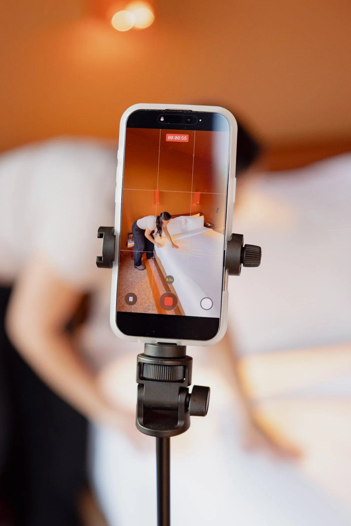 A person is recording a woman making a bed on a smartphone mounted on a tripod.