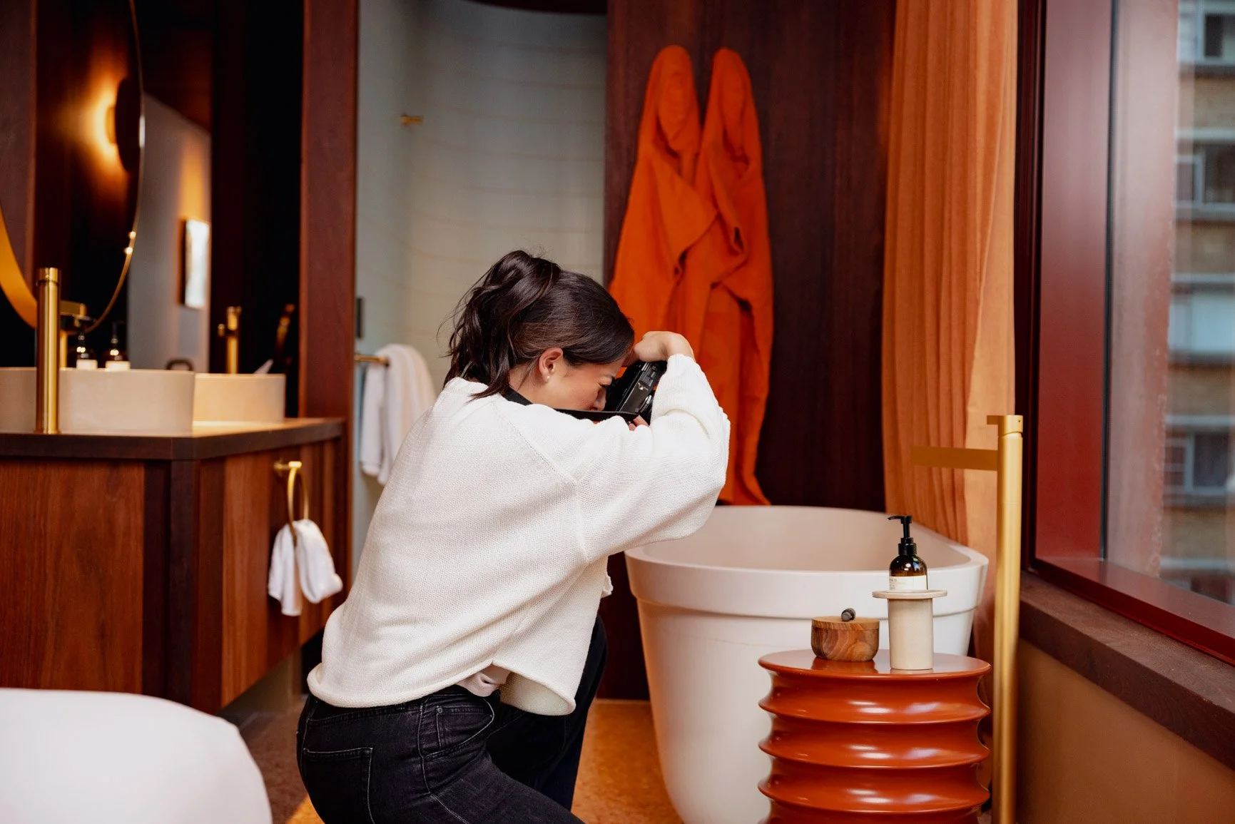A woman with short dark hair crouches by a bathroom sink, looking through a camera she is holding up to her face.