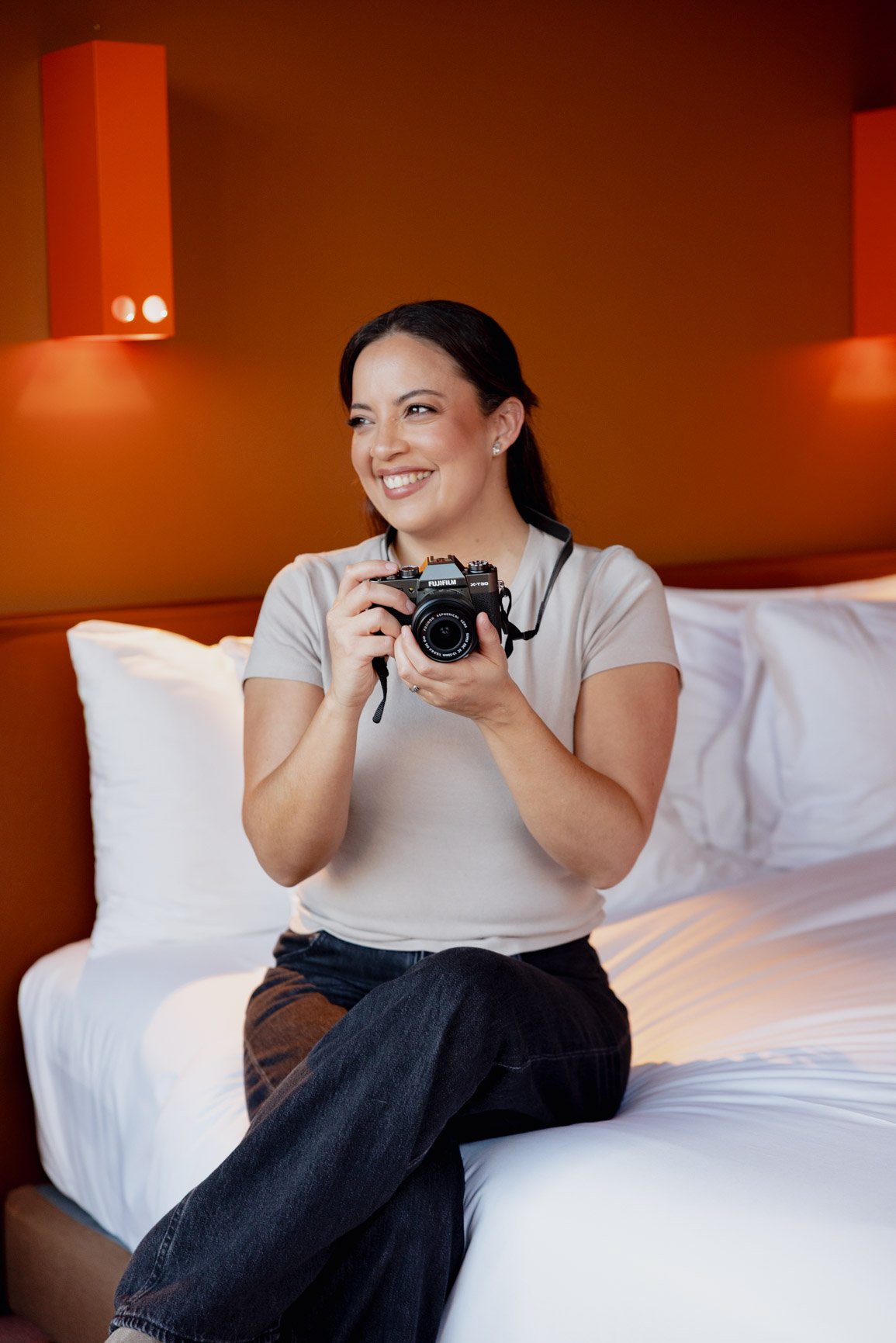 A woman sitting on a bed holding a camera, smiling, with a brown wall and wall-mounted lights in the background.