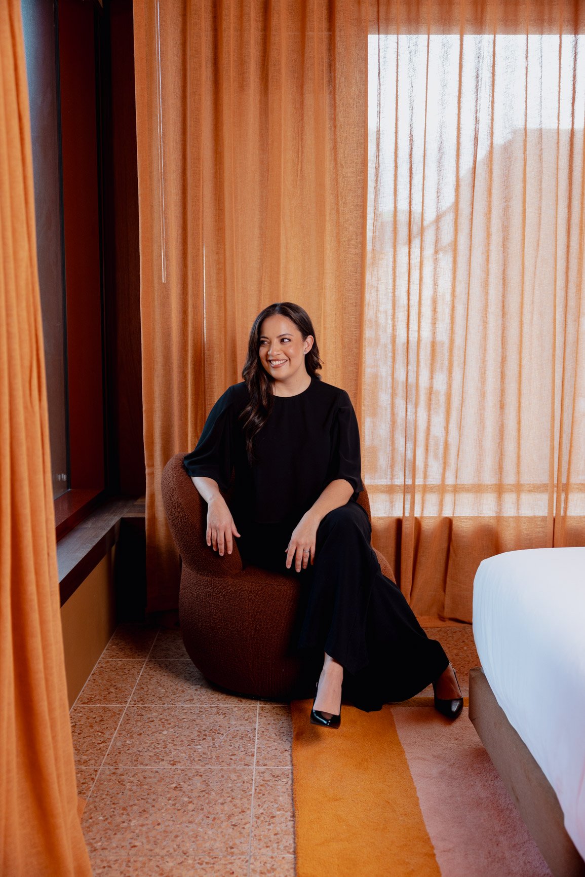 A woman sitting in an orange armchair with orange curtains behind her, wearing a black outfit and black high heels, smiling and looking to her left in a warmly lit room.