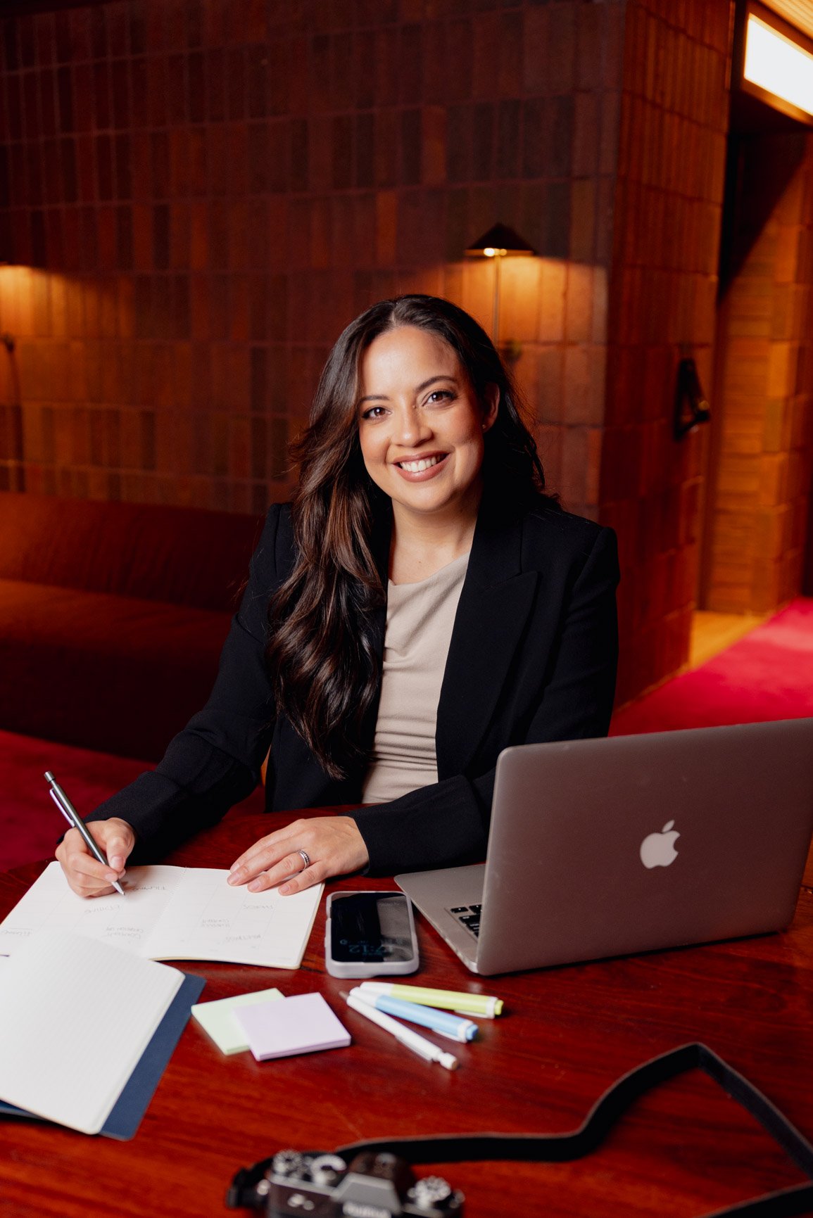 A smiling woman with long dark hair wearing a black blazer and beige top, sitting at a desk with a laptop, pen, notebooks, and stationery, in a warmly lit wood-paneled room.