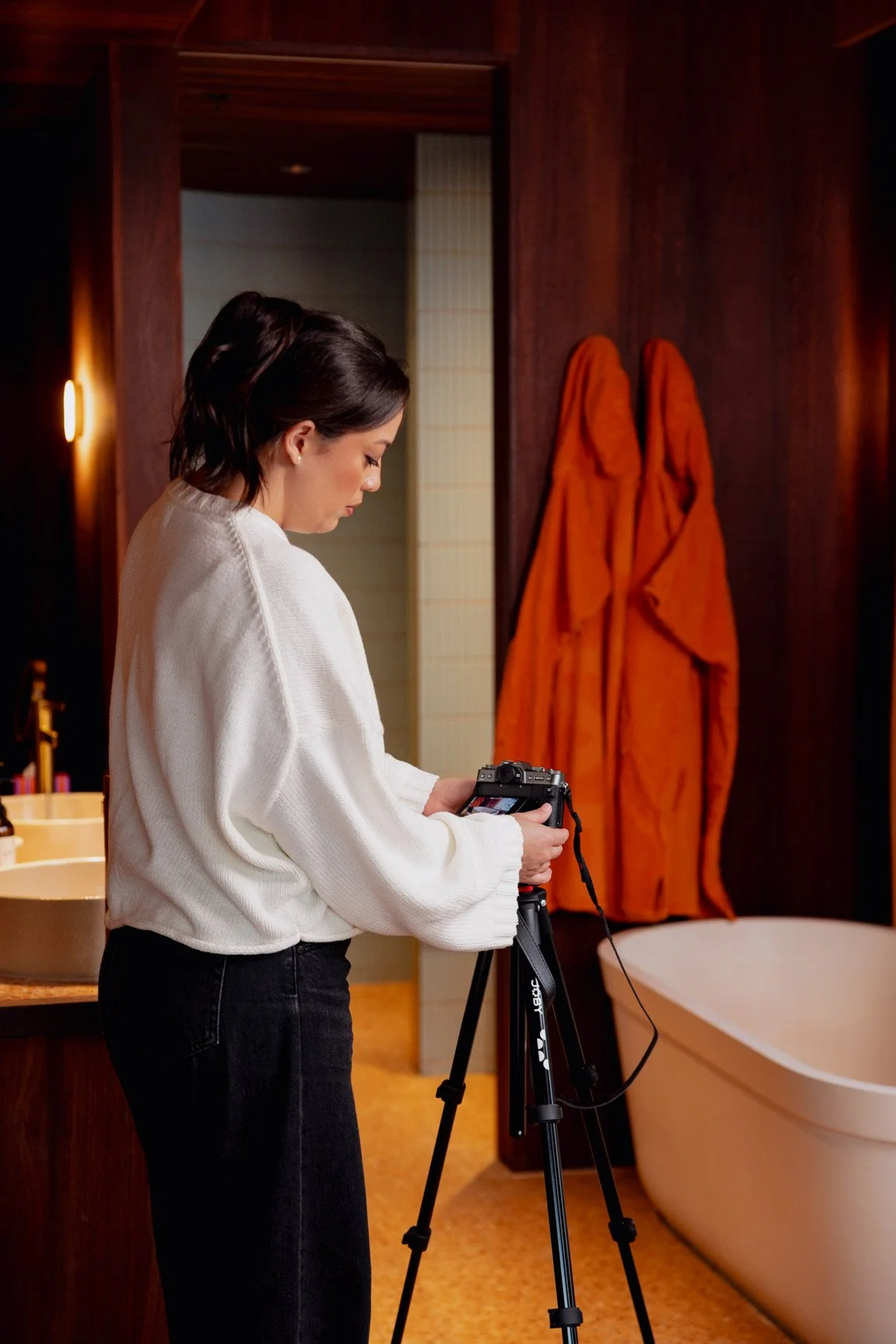 A woman with dark hair in a white sweater and black pants setting up a camera on a tripod in a hotel bathroom. Behind her are two orange robes hanging on the wall and a large bathtub.