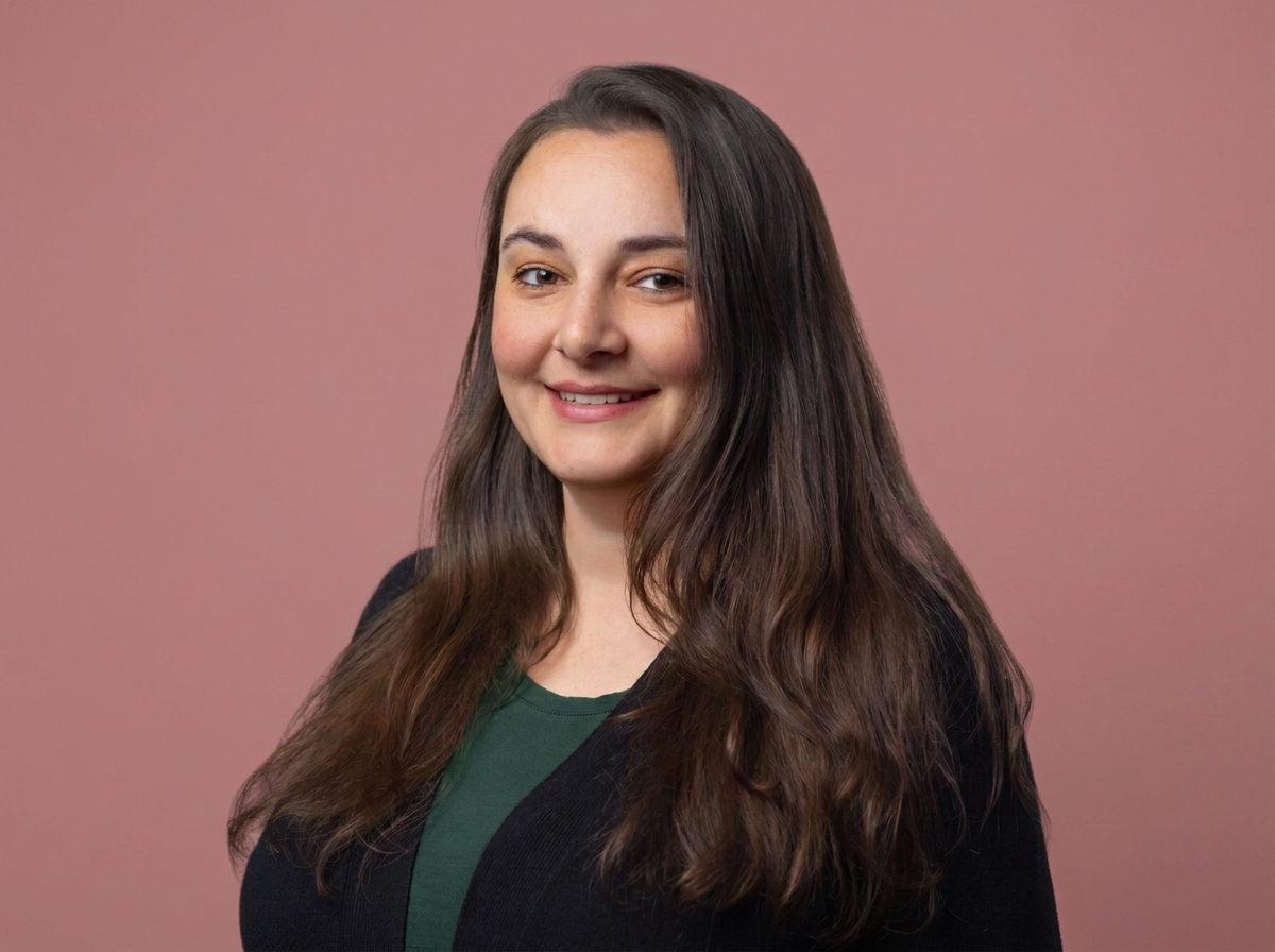 A woman with long brown hair wearing a black blazer and green top, smiling at the camera against a pink background.