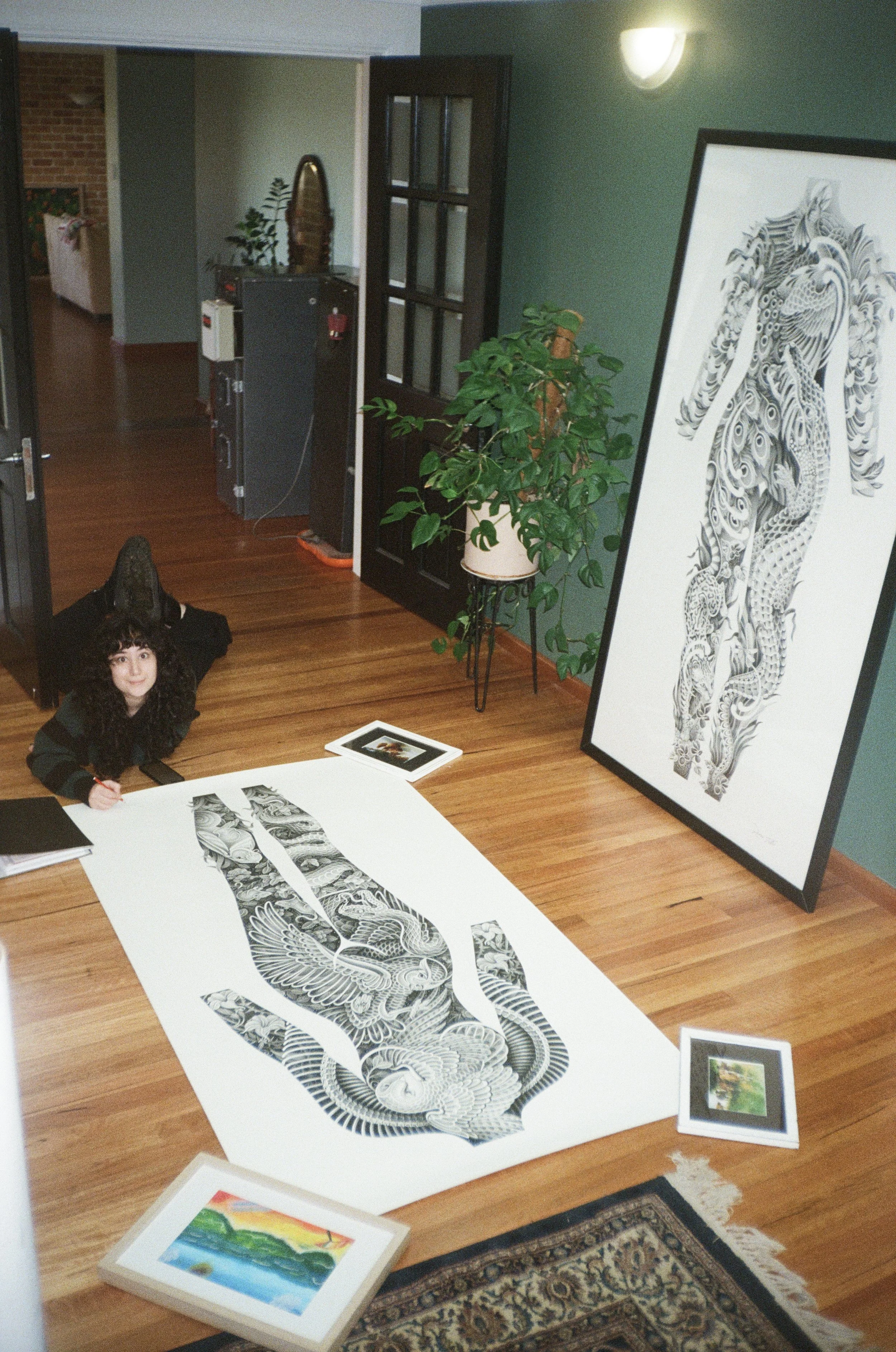 A person with curly dark hair and a black sweater is drawing or sketching artwork on a large sheet of paper on the floor. Around her, there are framed artworks, including a colorful landscape and a detailed black-and-white illustration of a bird with