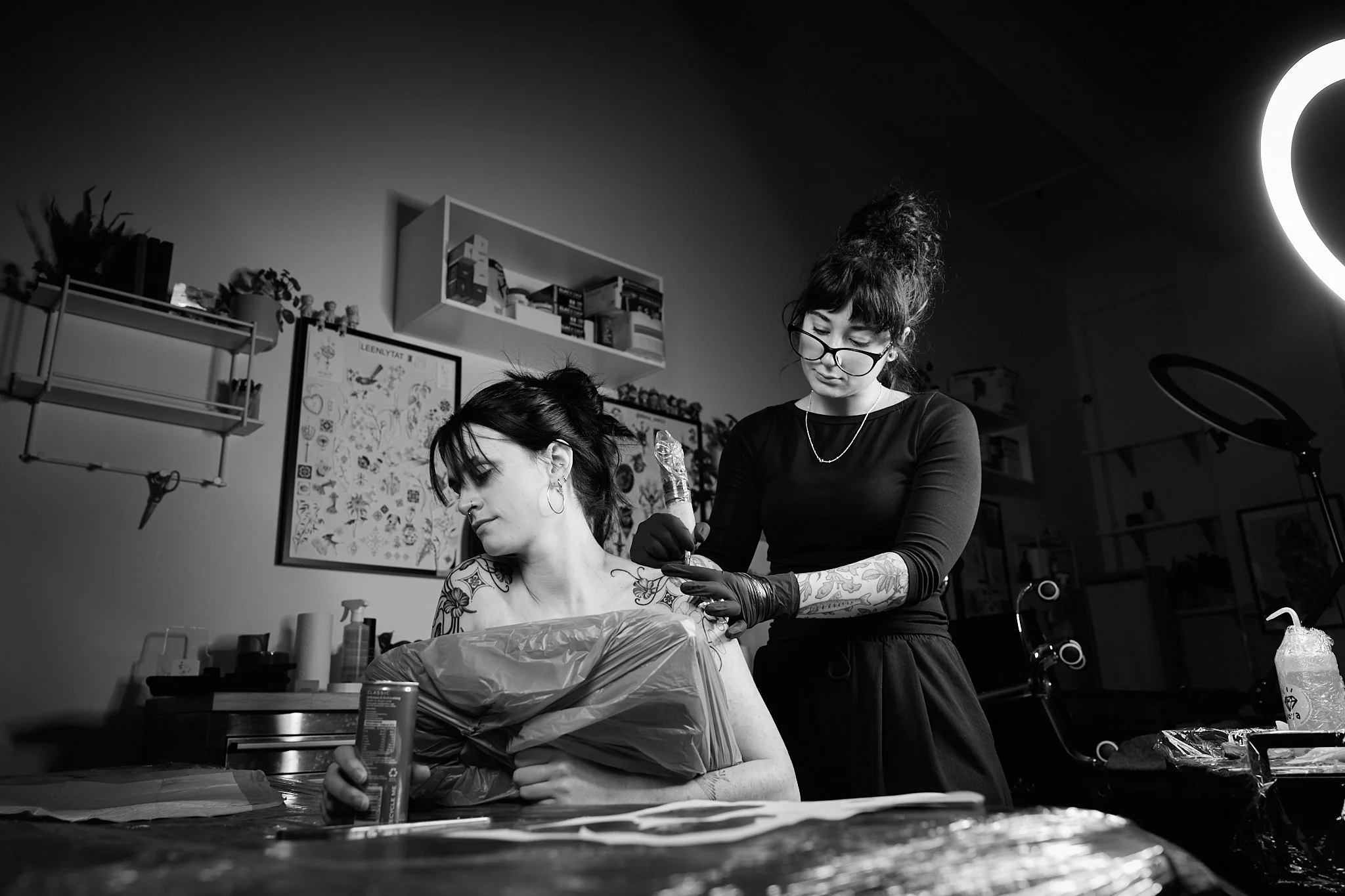 A tattoo artist is applying a tattoo to a person sitting at a table. The tattoo artist has glasses, tattoos on her arms, and is wearing gloves. The setting appears to be a tattoo studio with shelves, plants, and walls decorated with artwork.
