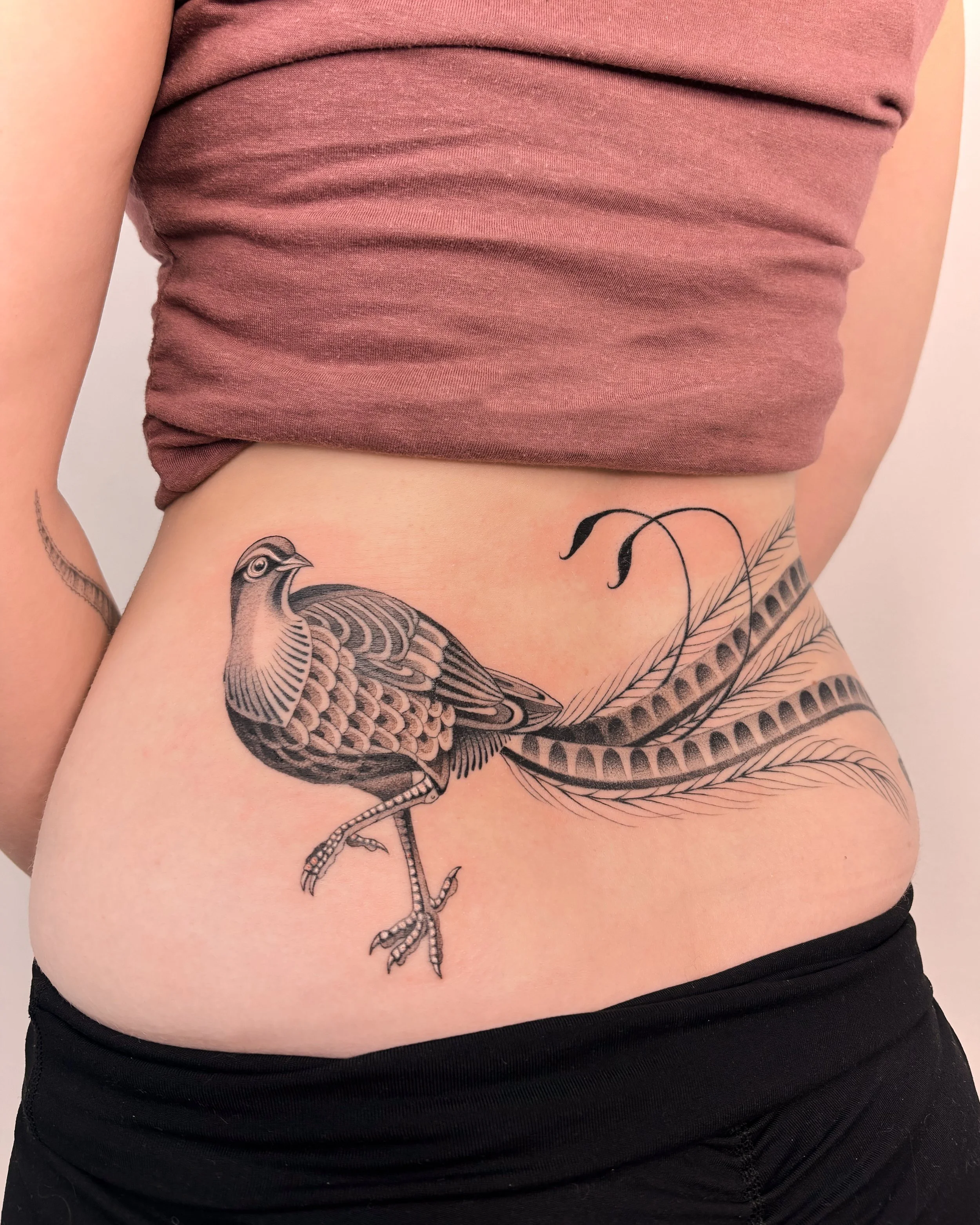 Tattoo of a bird perched on a branch on a person's side, partially visible, with a pink top and black pants.