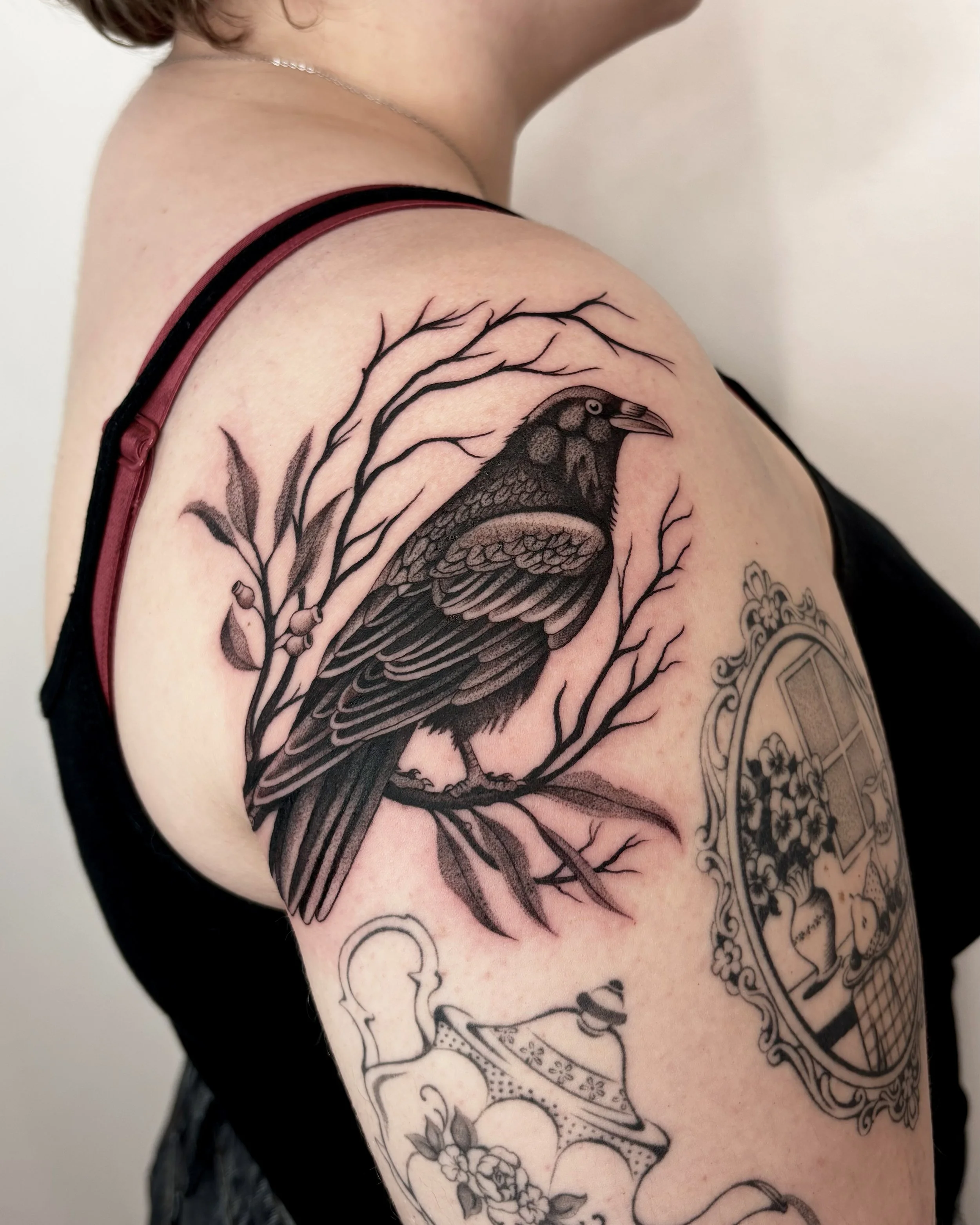 A close-up of a person's upper arm and shoulder featuring a black ink tattoo of a bird with intricate feathers, surrounded by branches with small berries and flowers. The person is wearing a dark red strap tank top and a silver necklace.
