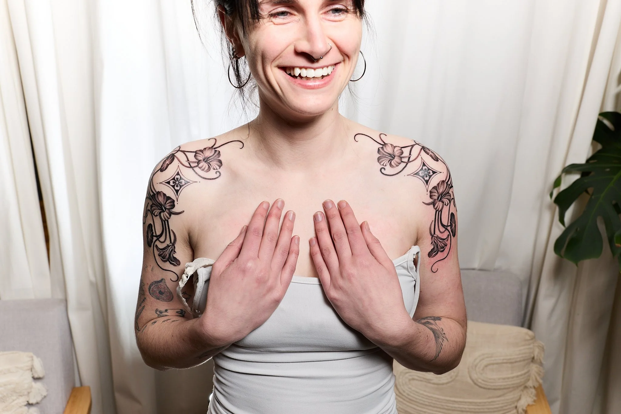 A smiling woman with tattoos on her shoulders and arms, holding her hands over her chest, standing in front of light-colored curtains and plants.