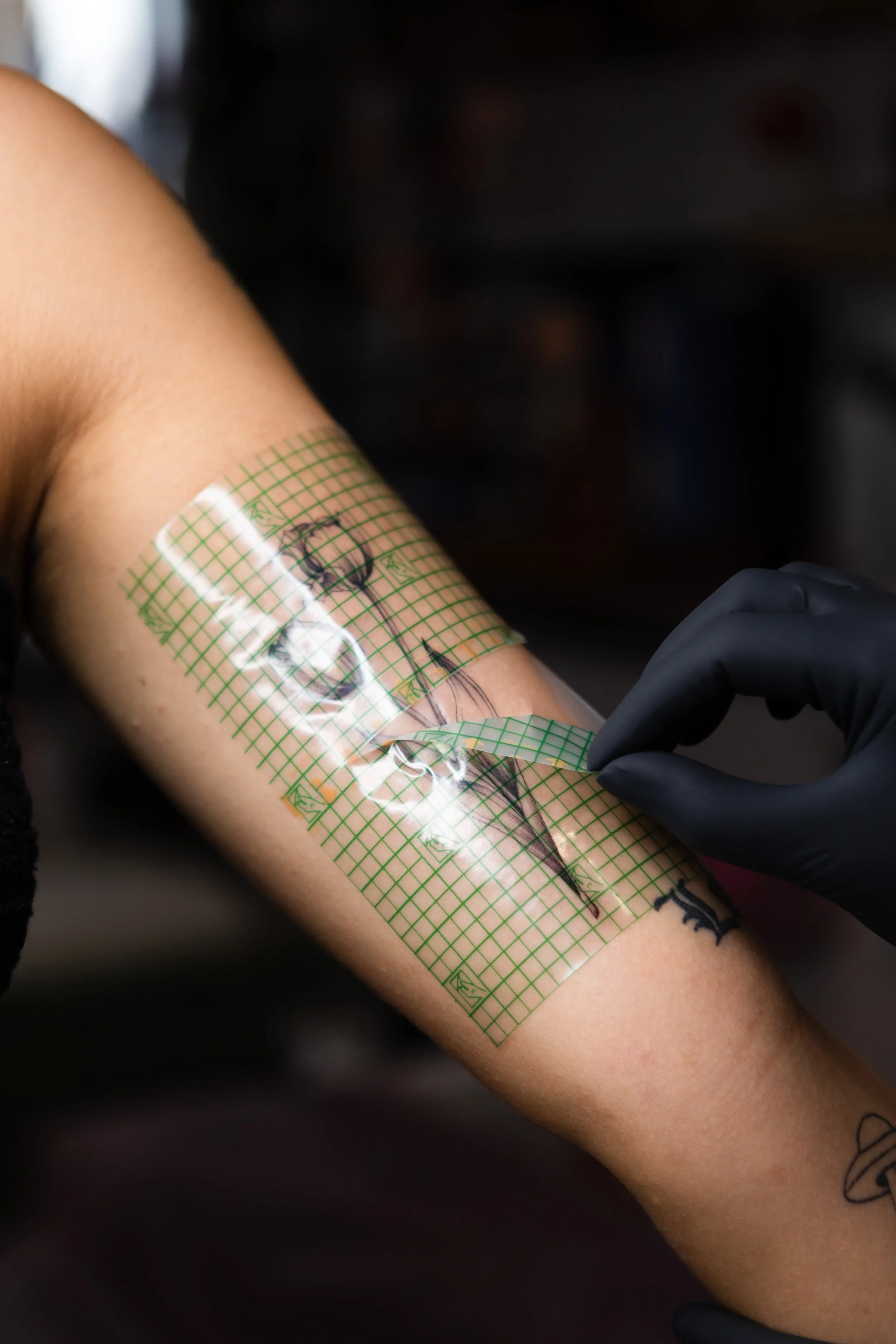 A person with tattoos is getting a tattoo on their arm. A tattoo artist with black gloves is working on the tattoo.