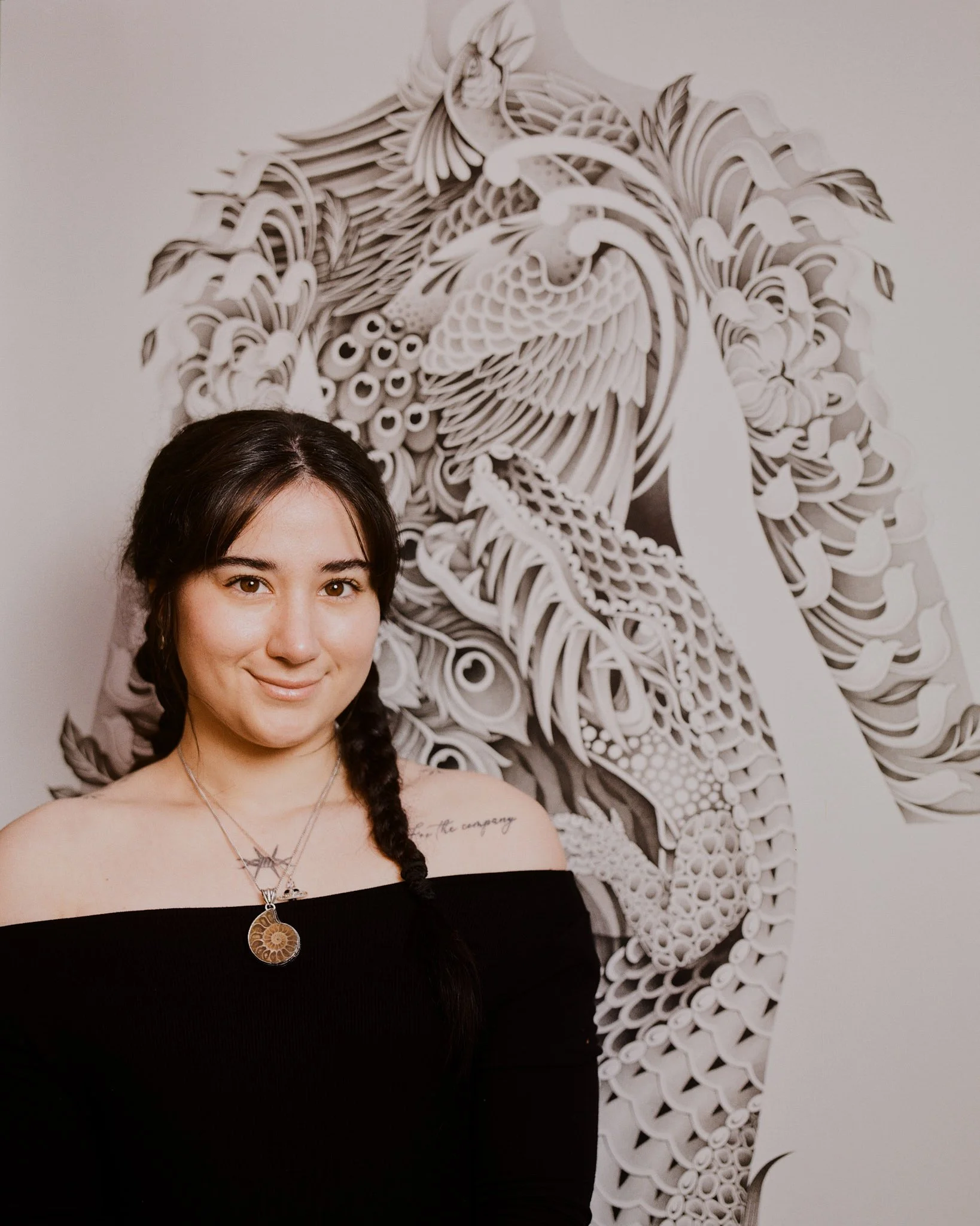A woman with dark hair and brown eyes smiling, wearing a black off-the-shoulder top, jewelry, and a tattoo on her collarbone, standing in front of a black-and-white paper cutout of a bird with detailed feathers on a plain wall.