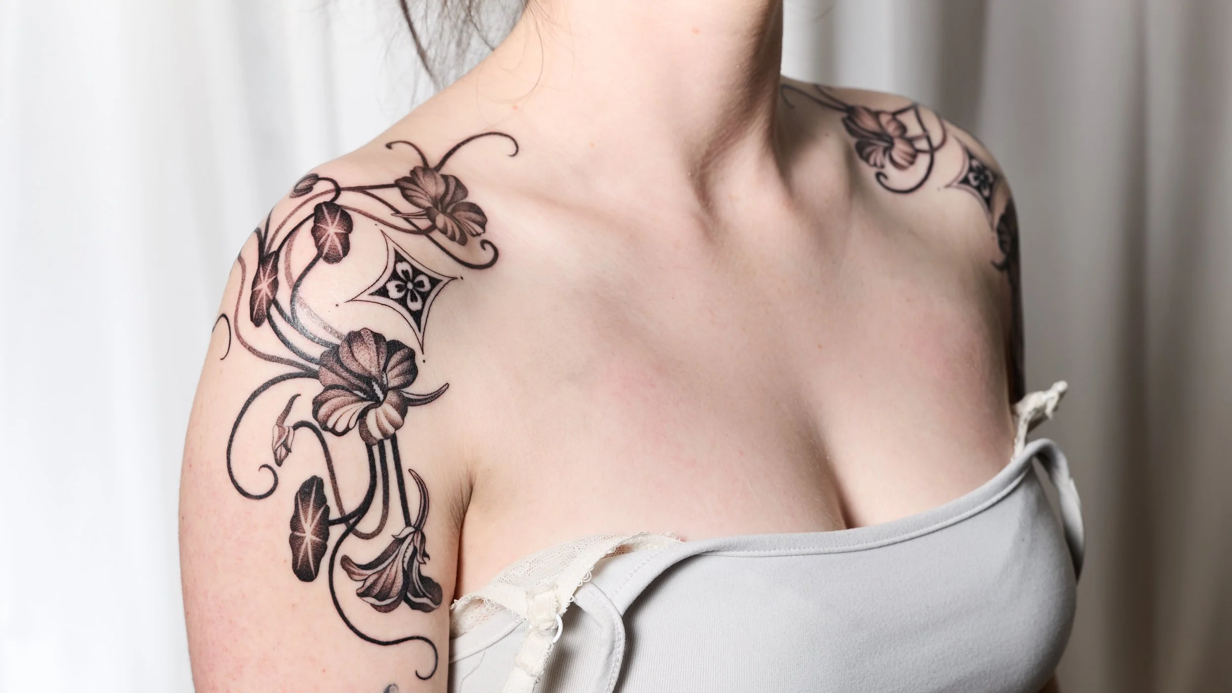 Close-up of a woman's shoulder and upper arm, showcasing a detailed black tattoo of flowers, leaves, and decorative patterns.