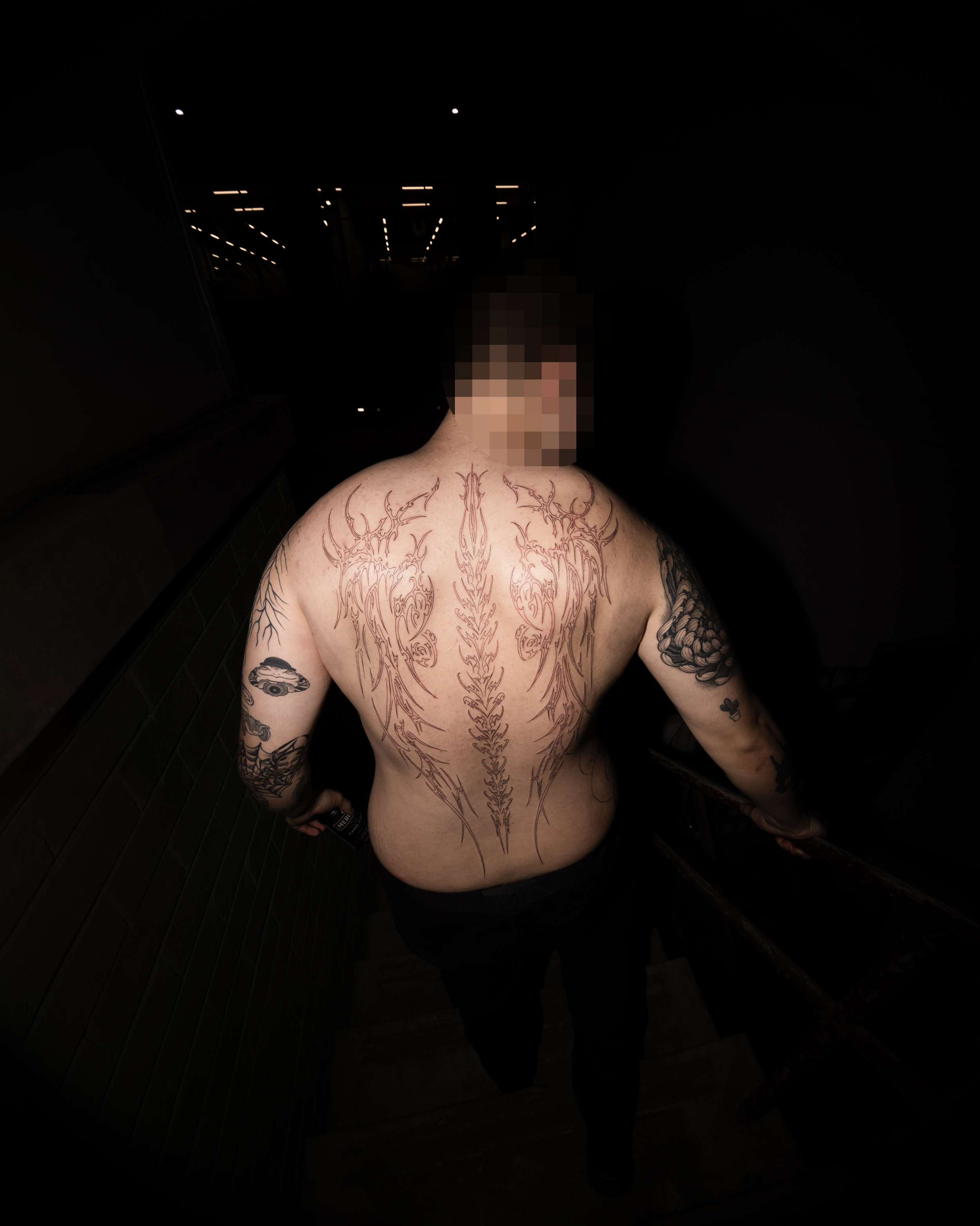 A shirtless man with tattoos on his back and arms, standing in a dark indoor space with ceiling lights reflected on the ceiling above.