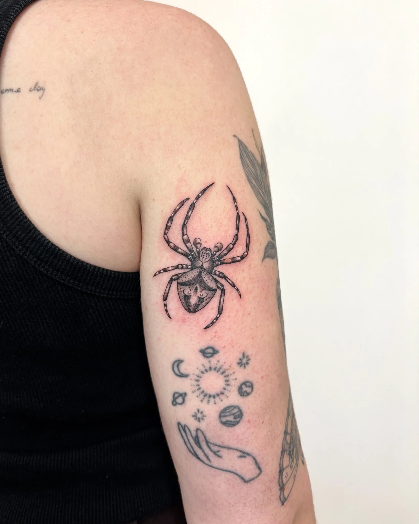 Close-up of a person’s upper arm with multiple tattoos, including a large black spider on the upper arm, and smaller tattoos of moons, planets, and celestial symbols on the forearm. The person is wearing a black sleeveless top.