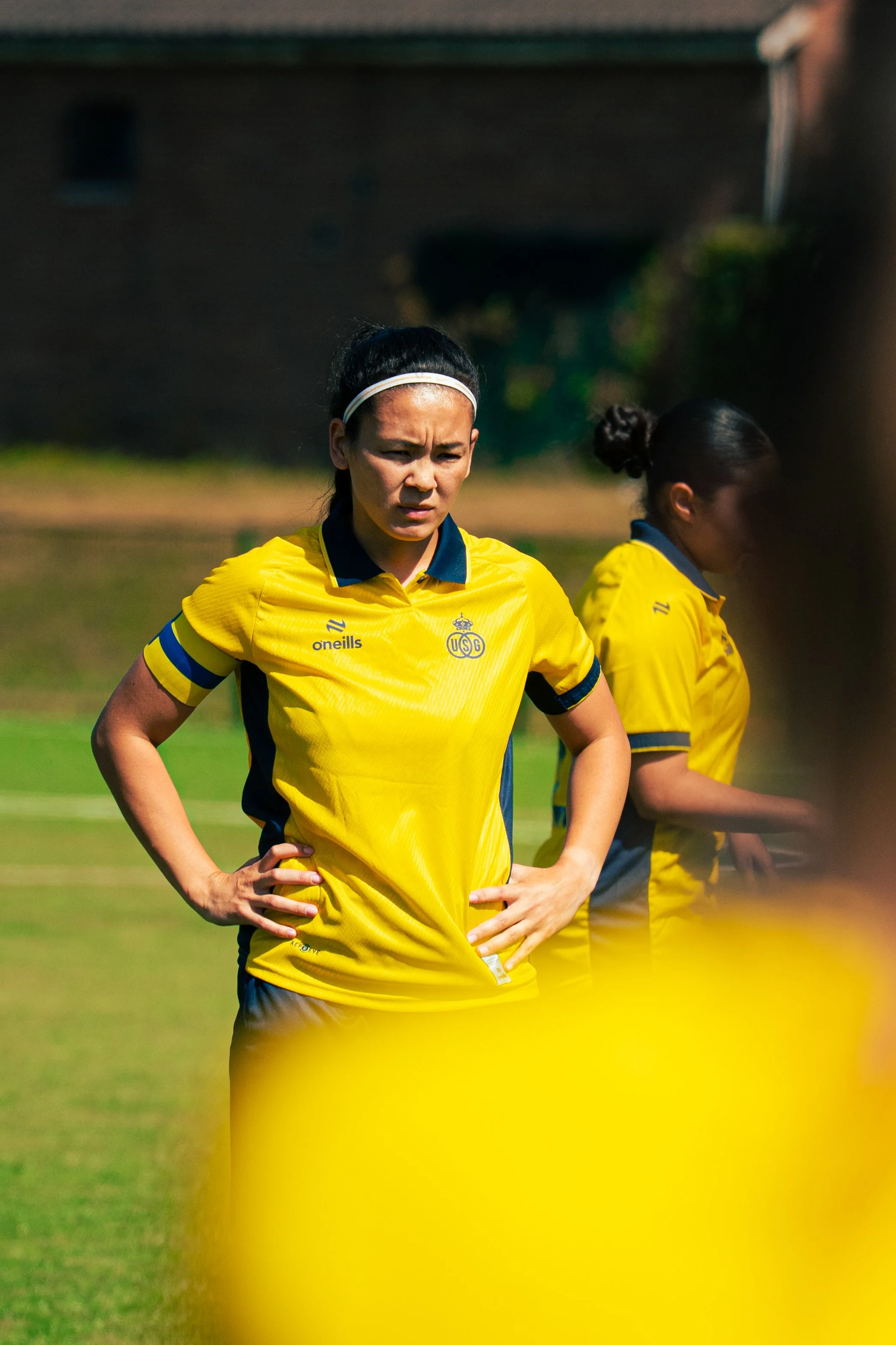 Female soccer player in yellow jersey with hand on hip, standing on grass field, focused expression.