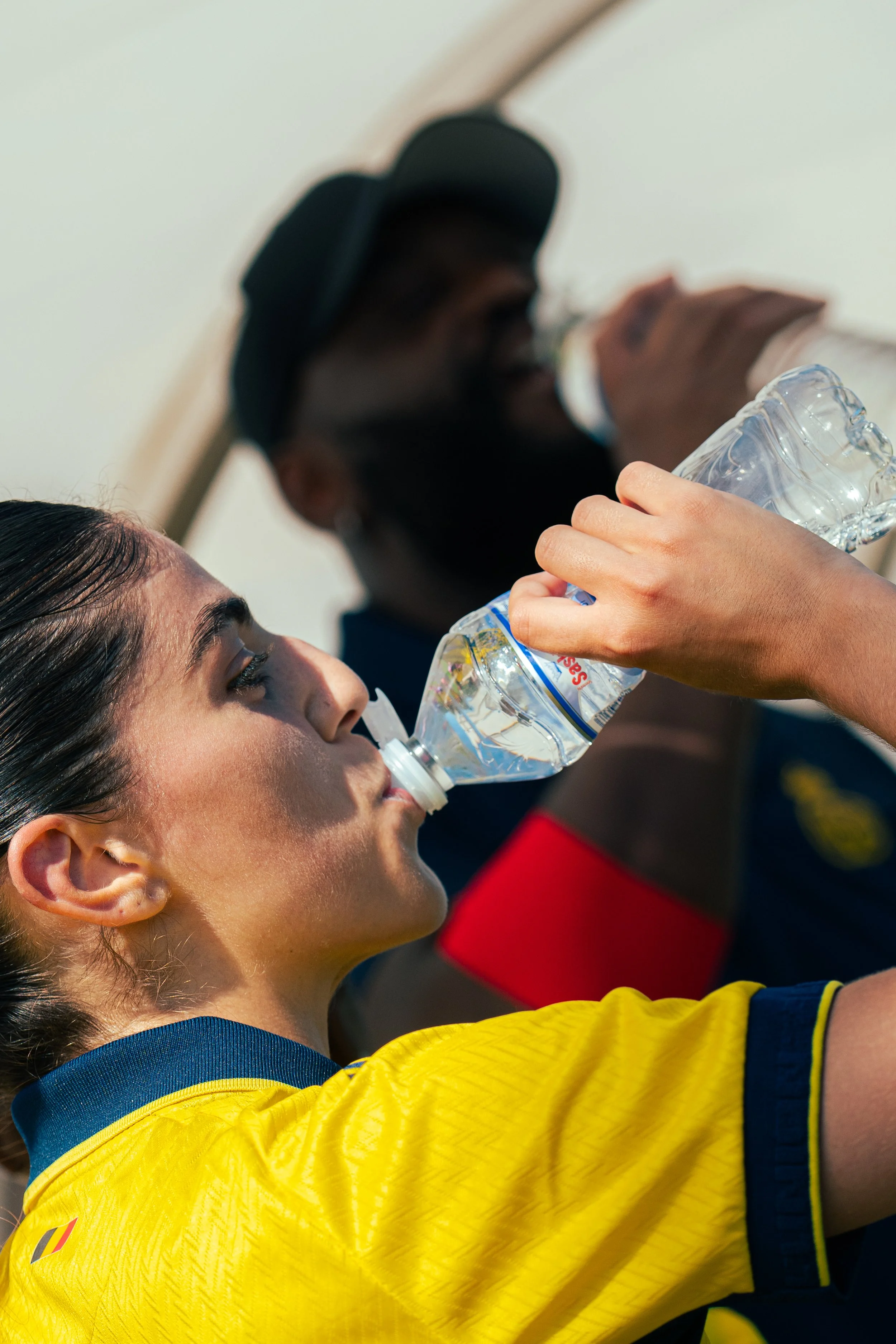 A young woman in a yellow and blue sports jersey drinking water from a clear plastic bottle, with a man in the background wearing a cap and face mask.