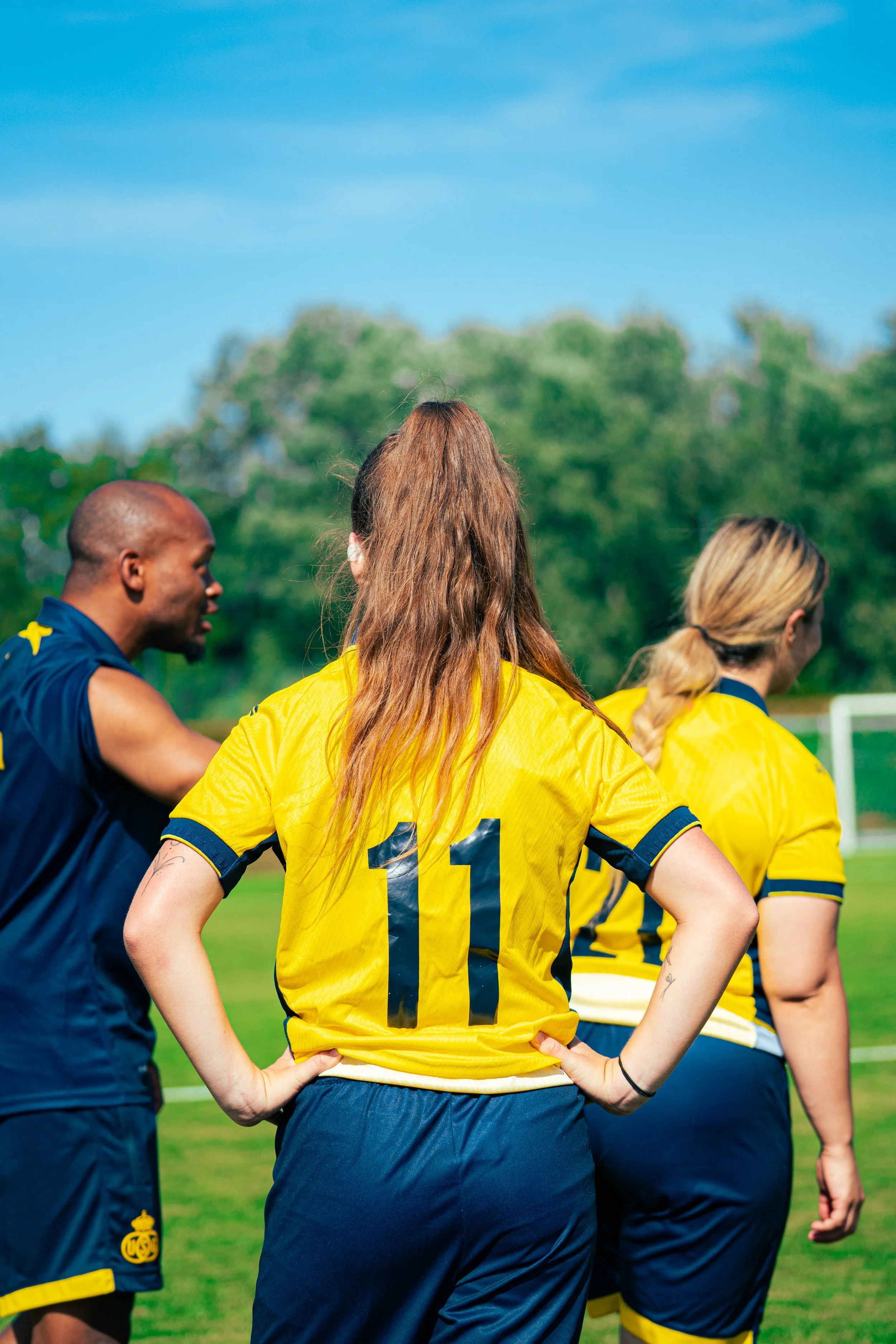 A group of female soccer players in yellow jerseys gathered on the field during daytime. One player has the number 11 on her jersey. They are standing with their backs facing the camera, with a background of green trees and a blue sky.