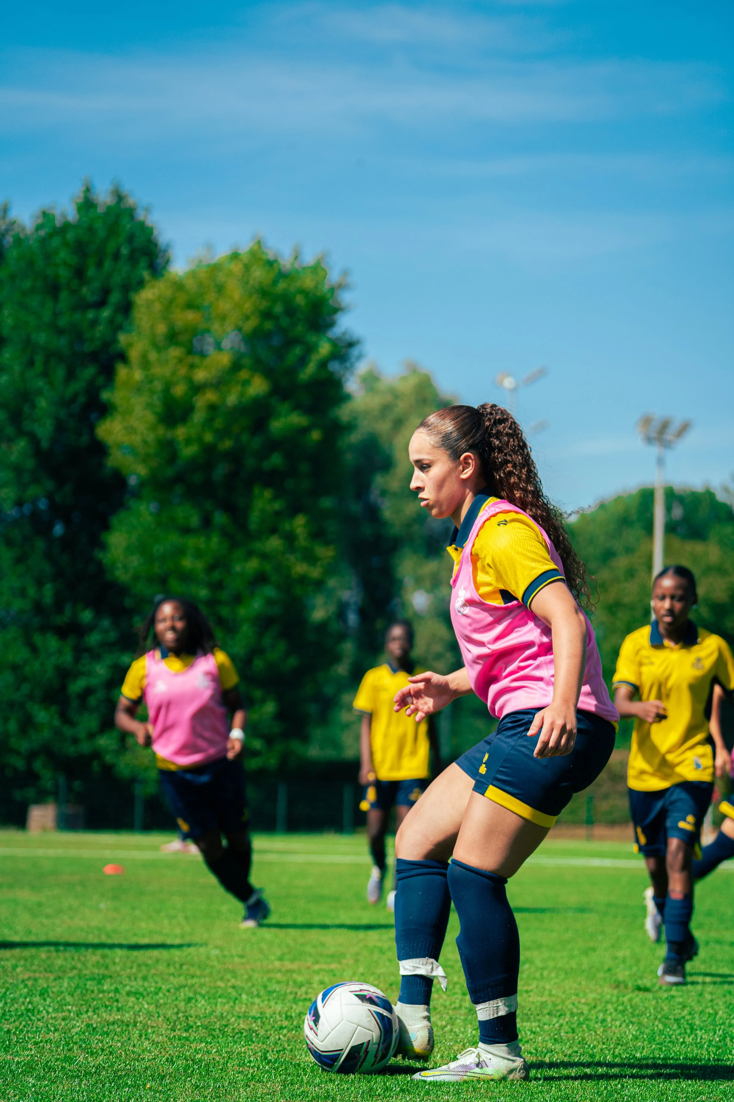 A female soccer player with curly hair, wearing a yellow and pink uniform, is about to kick a soccer ball on a field. Several other players in similar uniforms are running in the background, with trees and a blue sky overhead.