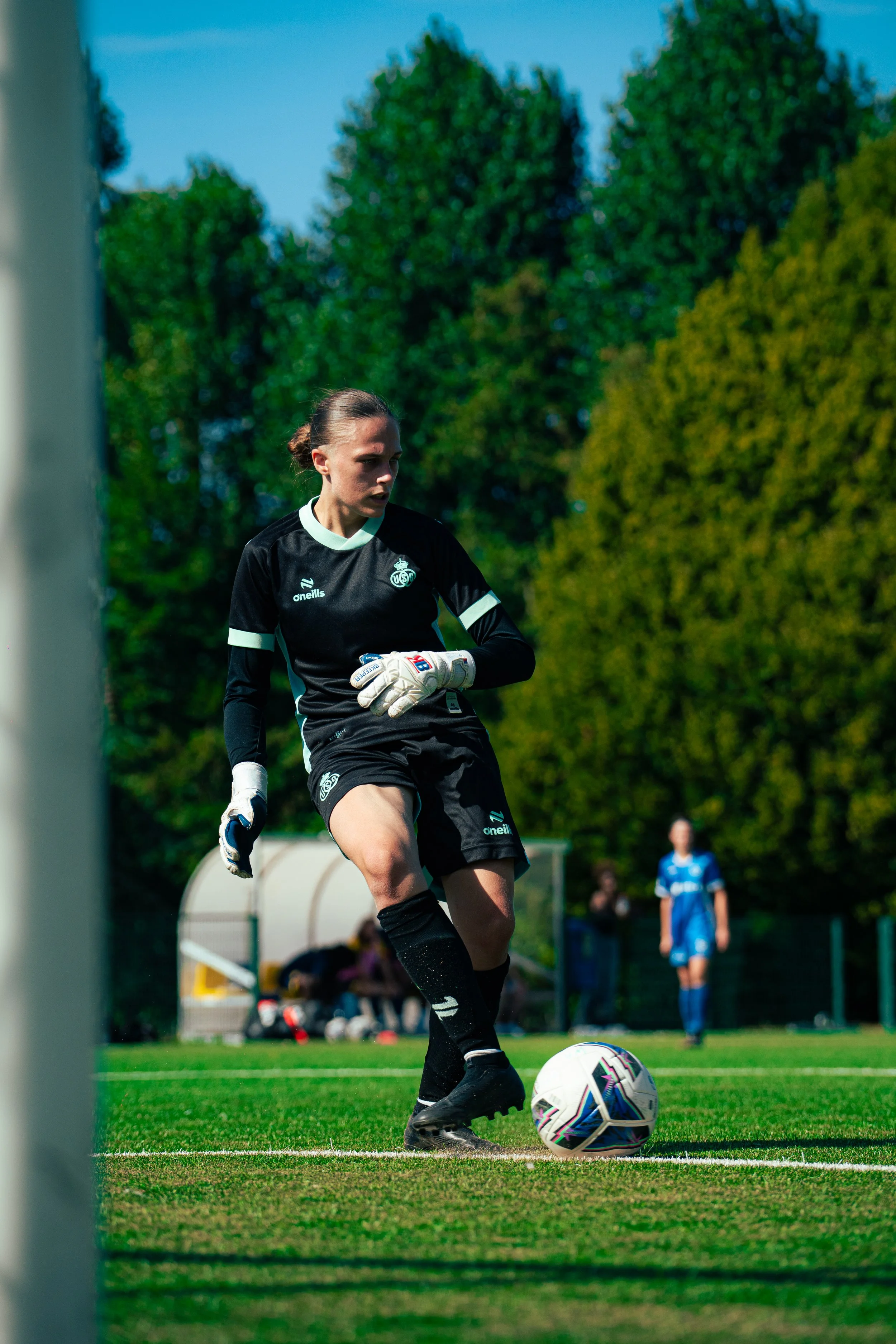 Female soccer player in black uniform, wearing goalkeeper gloves, preparing to kick a white soccer ball on the field during a game with green trees and other players in the background.