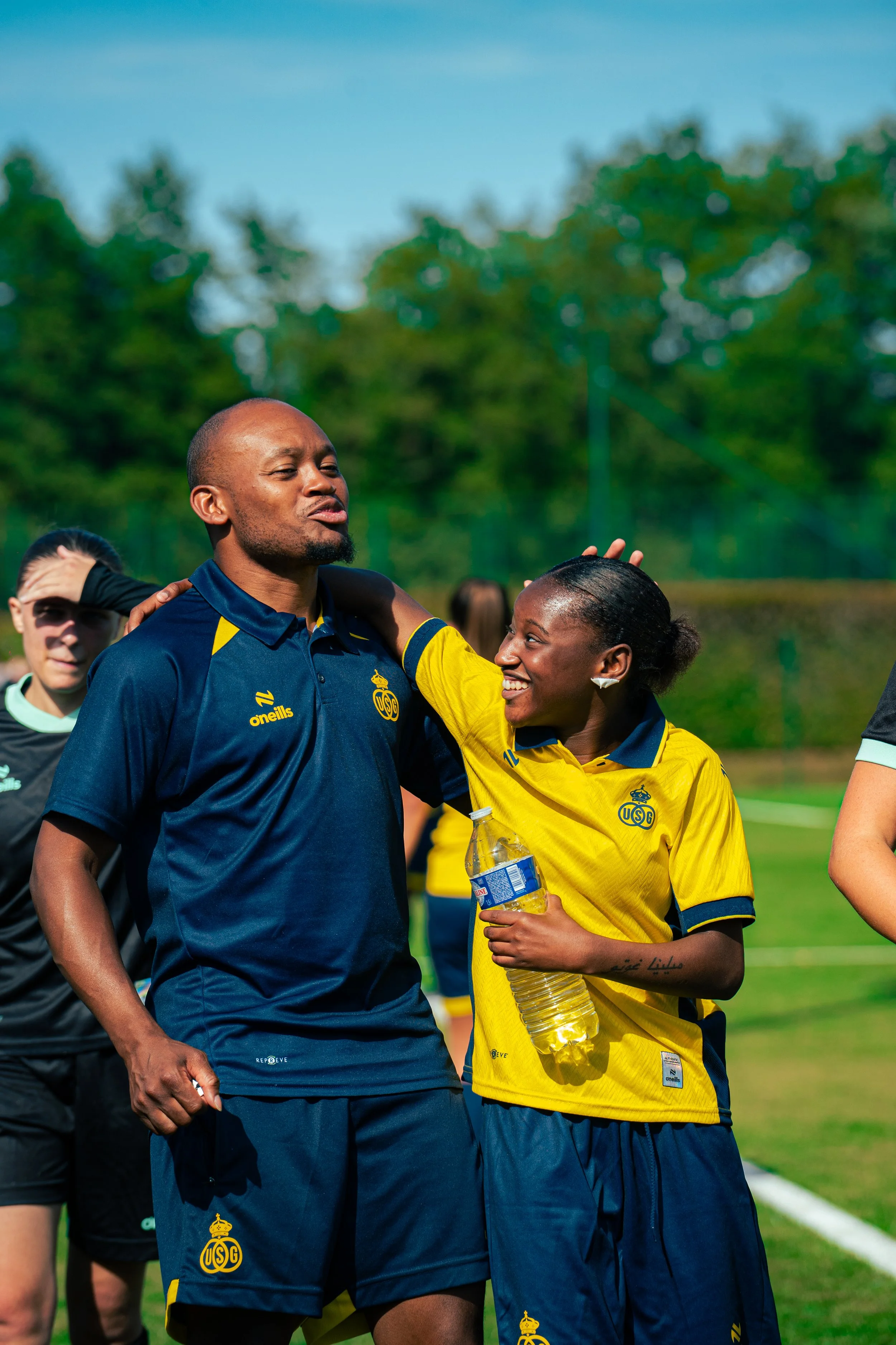 Two soccer players, a man and a woman, are smiling and talking on a soccer field. The man is wearing a navy blue jersey, and the woman is wearing a yellow jersey, holding a water bottle. They seem to be celebrating or sharing a moment after a game.