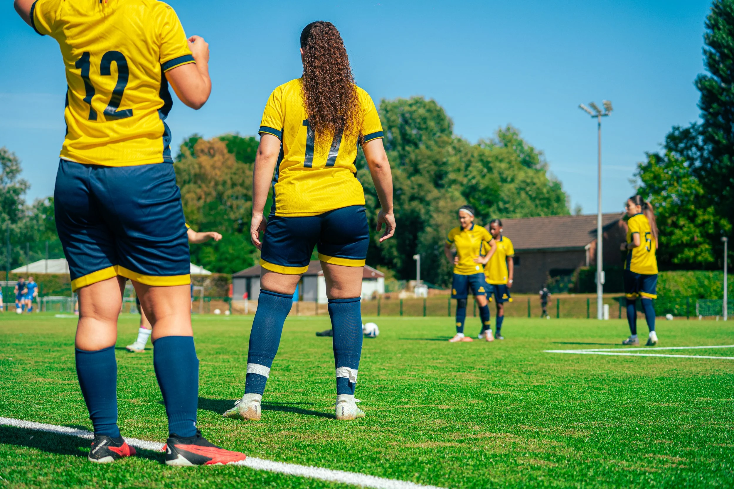 Soccer players in yellow jerseys standing on a grassy field, preparing for the game, with teammates in the background and trees under a blue sky.