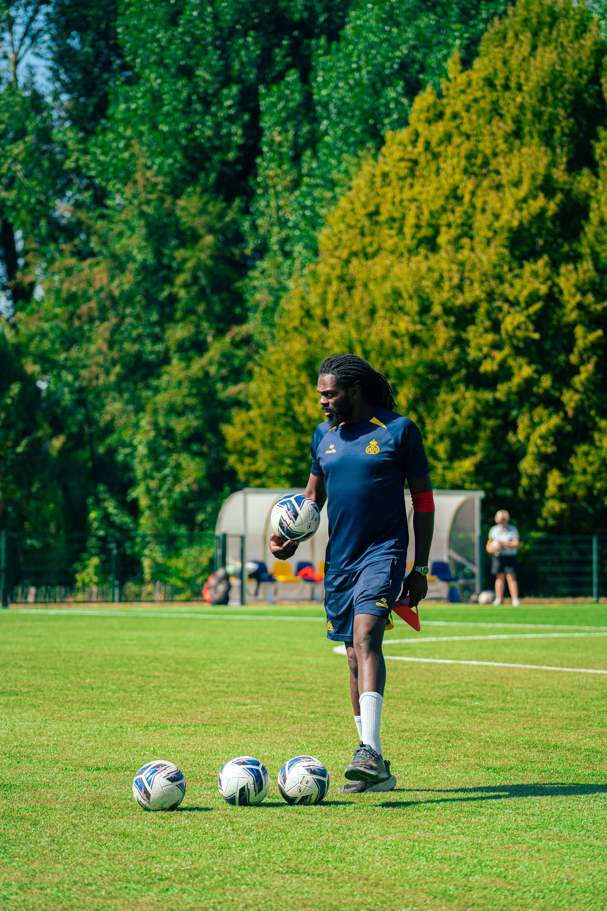 A man in a navy blue soccer uniform standing on a grassy field holding a soccer ball, surrounded by four more soccer balls on the ground, with trees and a person in the background.
