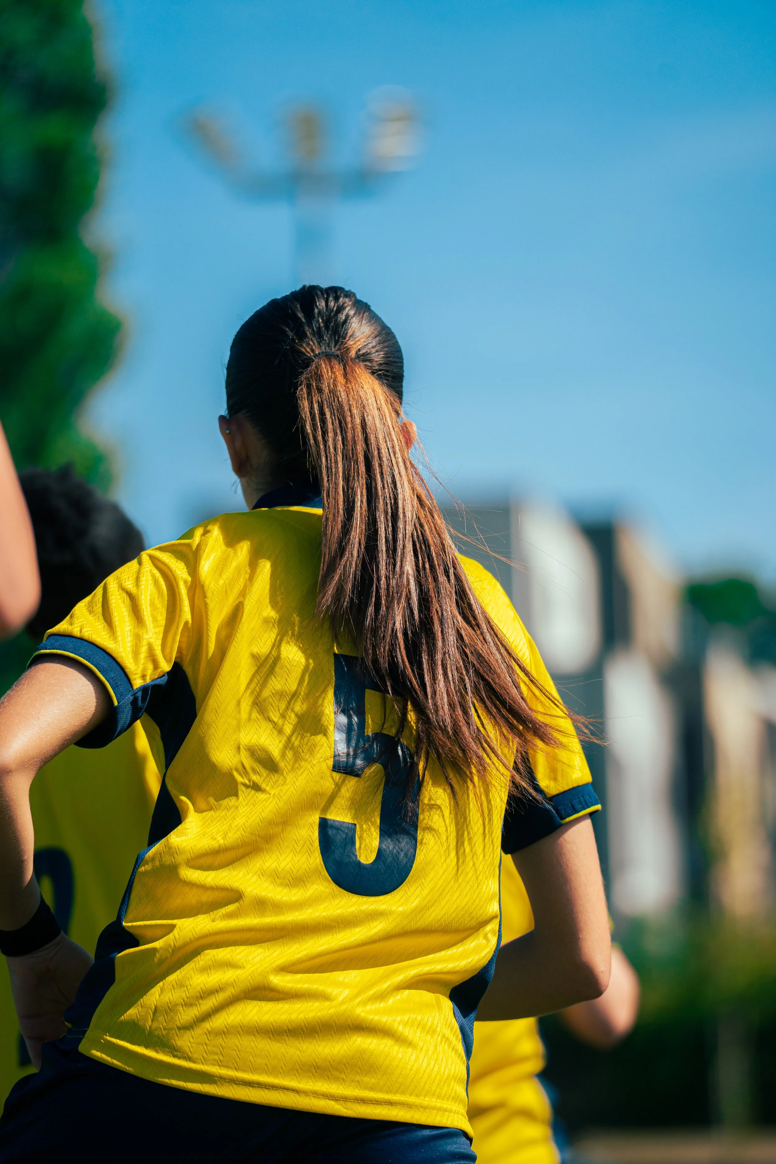 A female soccer player wearing a yellow jersey with the number 6 on the back, tied-back hair, and standing on a field during a game. There are other players and trees visible in the background.