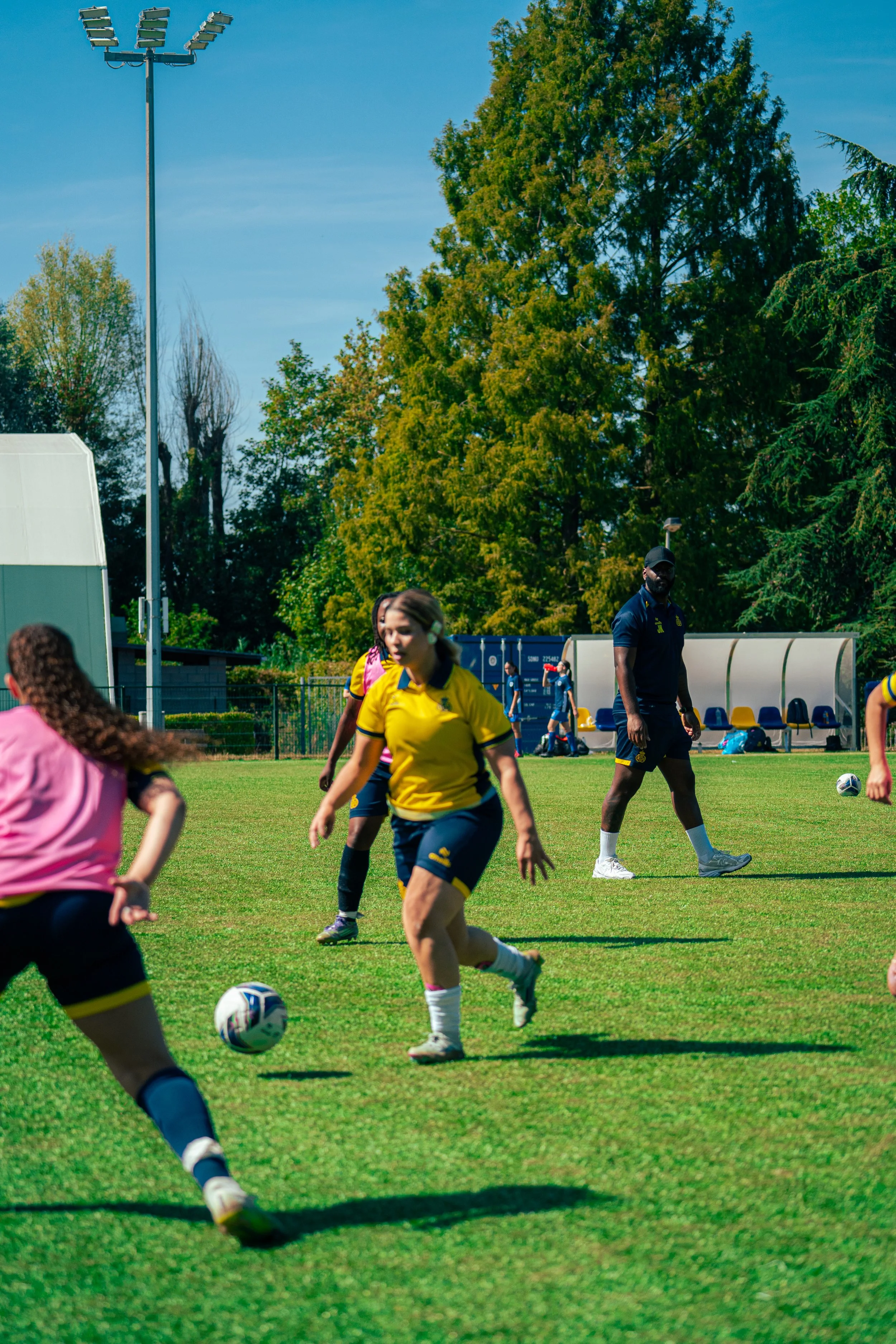 Women playing soccer on a green field during daytime, with a coach observing and large trees in the background.