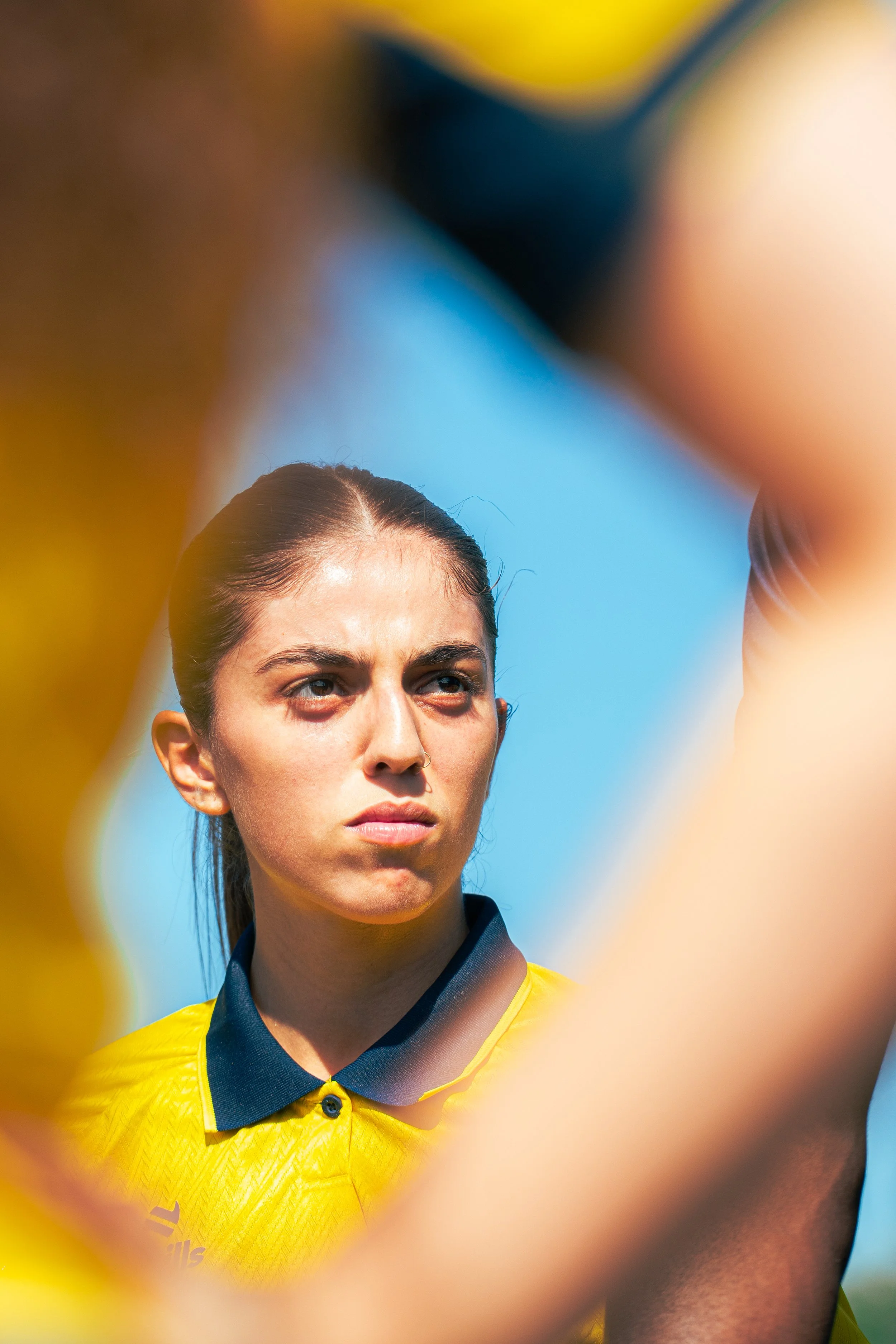 Close-up photo of a female soccer player with braided hair, wearing a yellow jersey, looking focused during a game with a blue sky in the background.