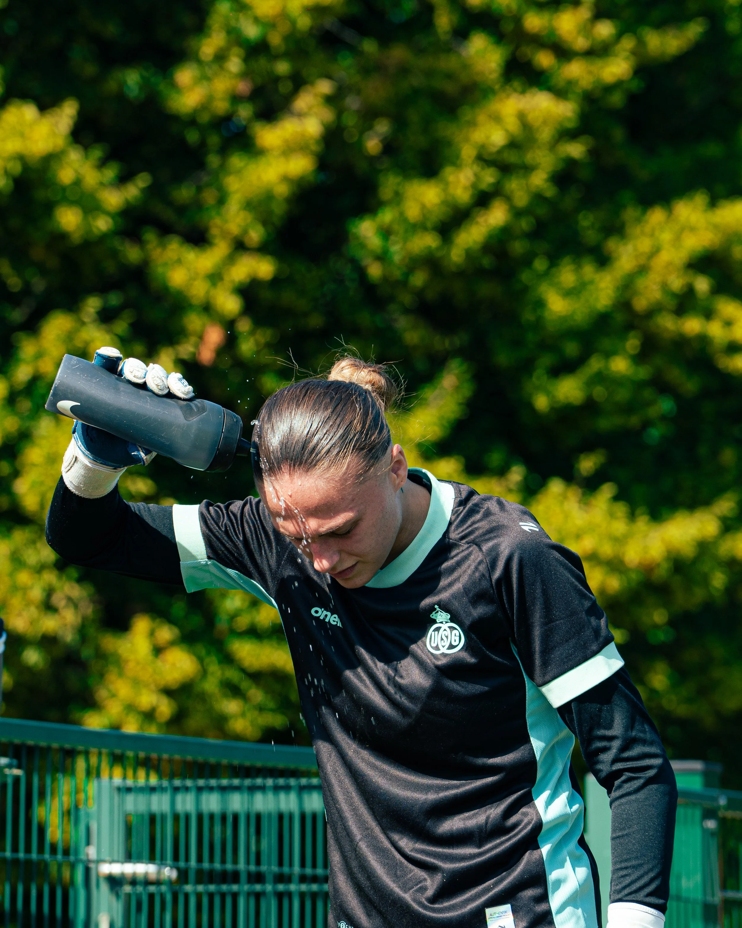 Close-up of a female tennis player with wet hair, wearing a black and light green tennis jersey, wiping sweat from her forehead with her hand, with a green fence and blurred trees in the background.
