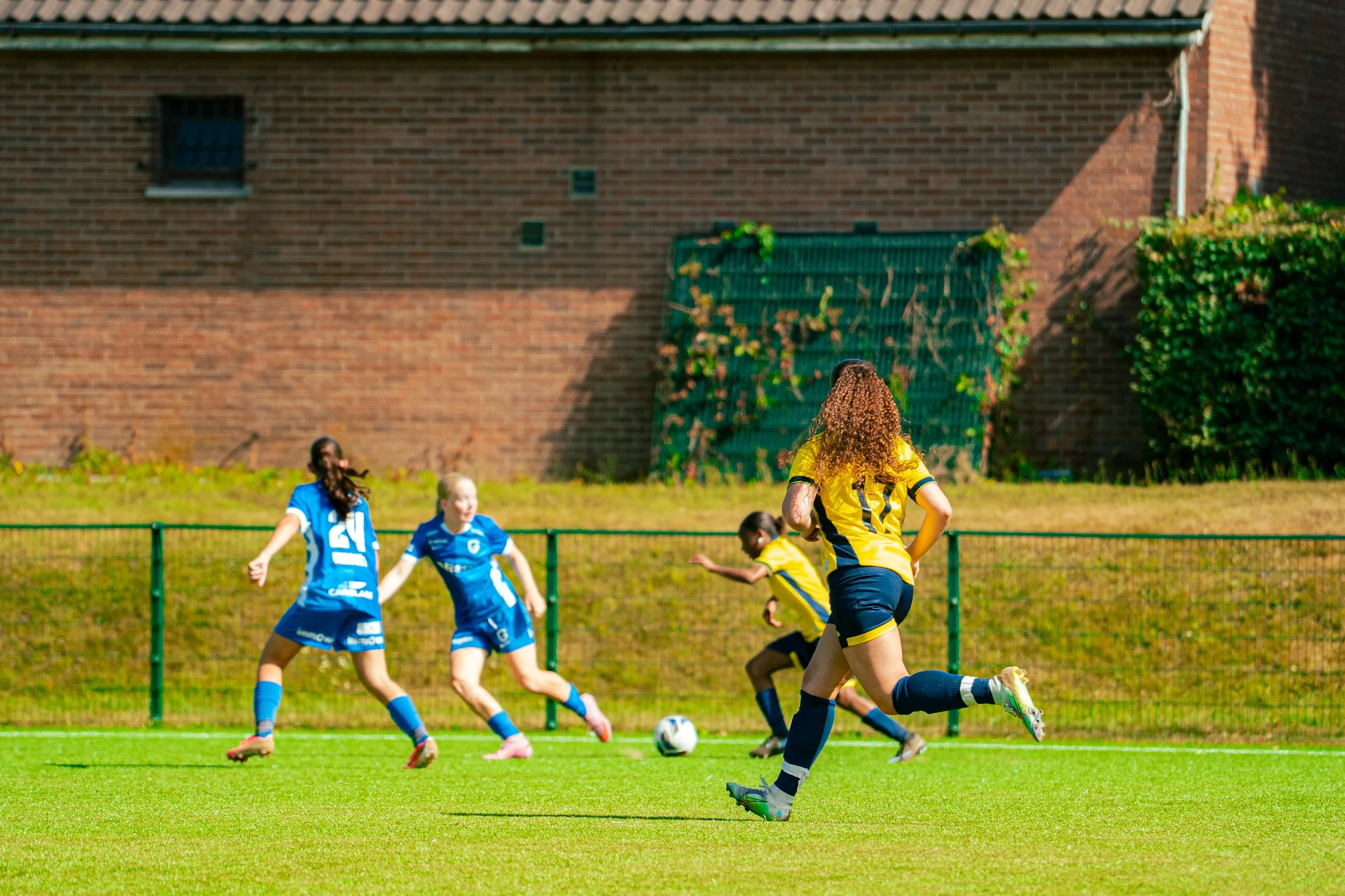 A women's soccer match in progress on a green field with a brick building in the background. Several players are running and chasing the soccer ball, with one player in a yellow and black uniform in the foreground and others in blue uniforms nearby.