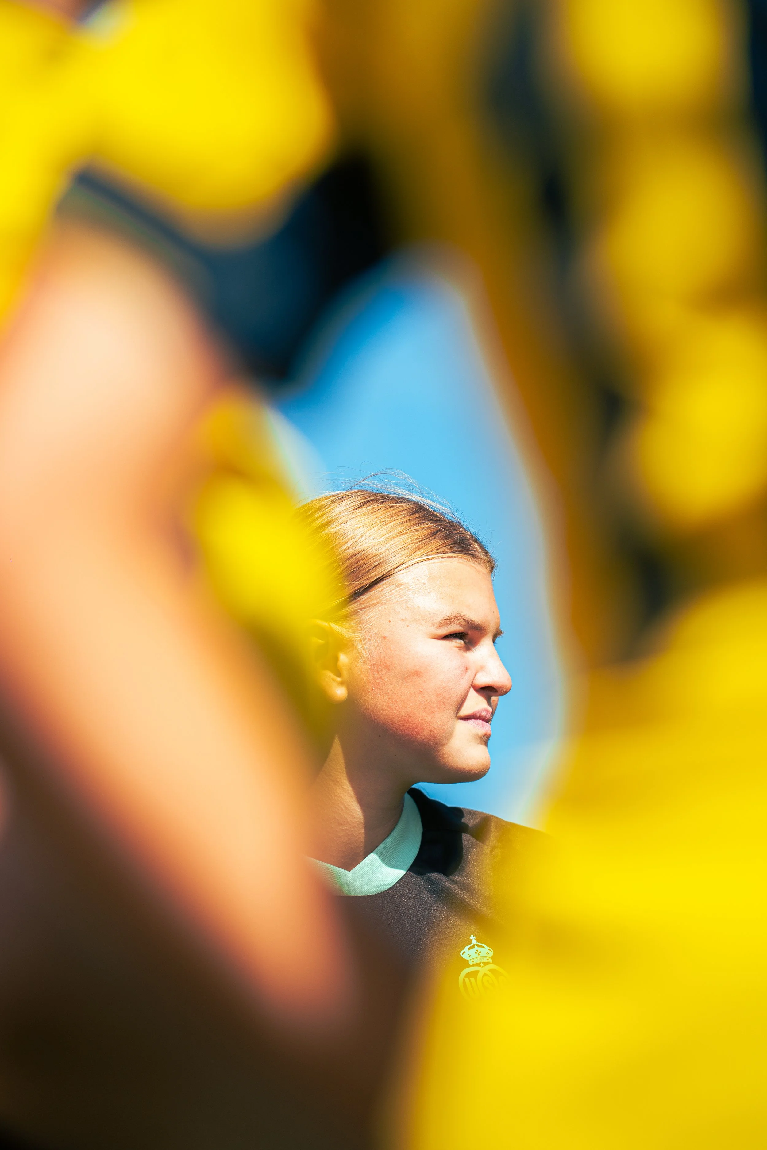 Close-up of a young woman with blonde hair smiling, wearing a black sports jersey, seen from the side, with blurred yellow and blue background.