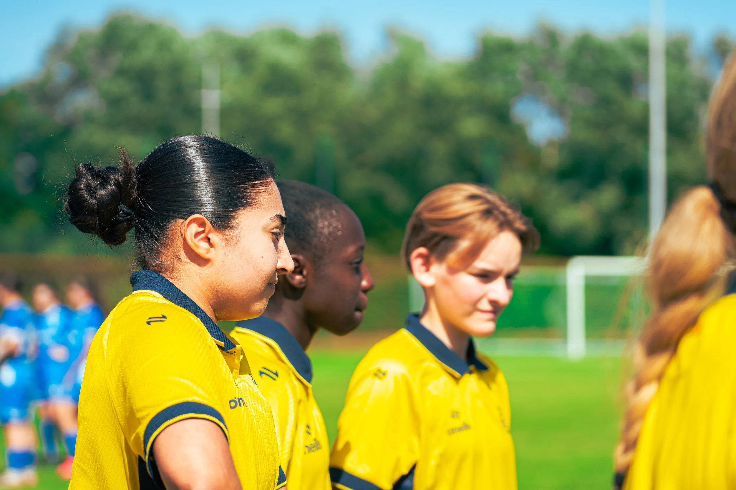 A group of young women standing in a line, wearing yellow sports jerseys, likely during a sports event on a sunny day with trees in the background.