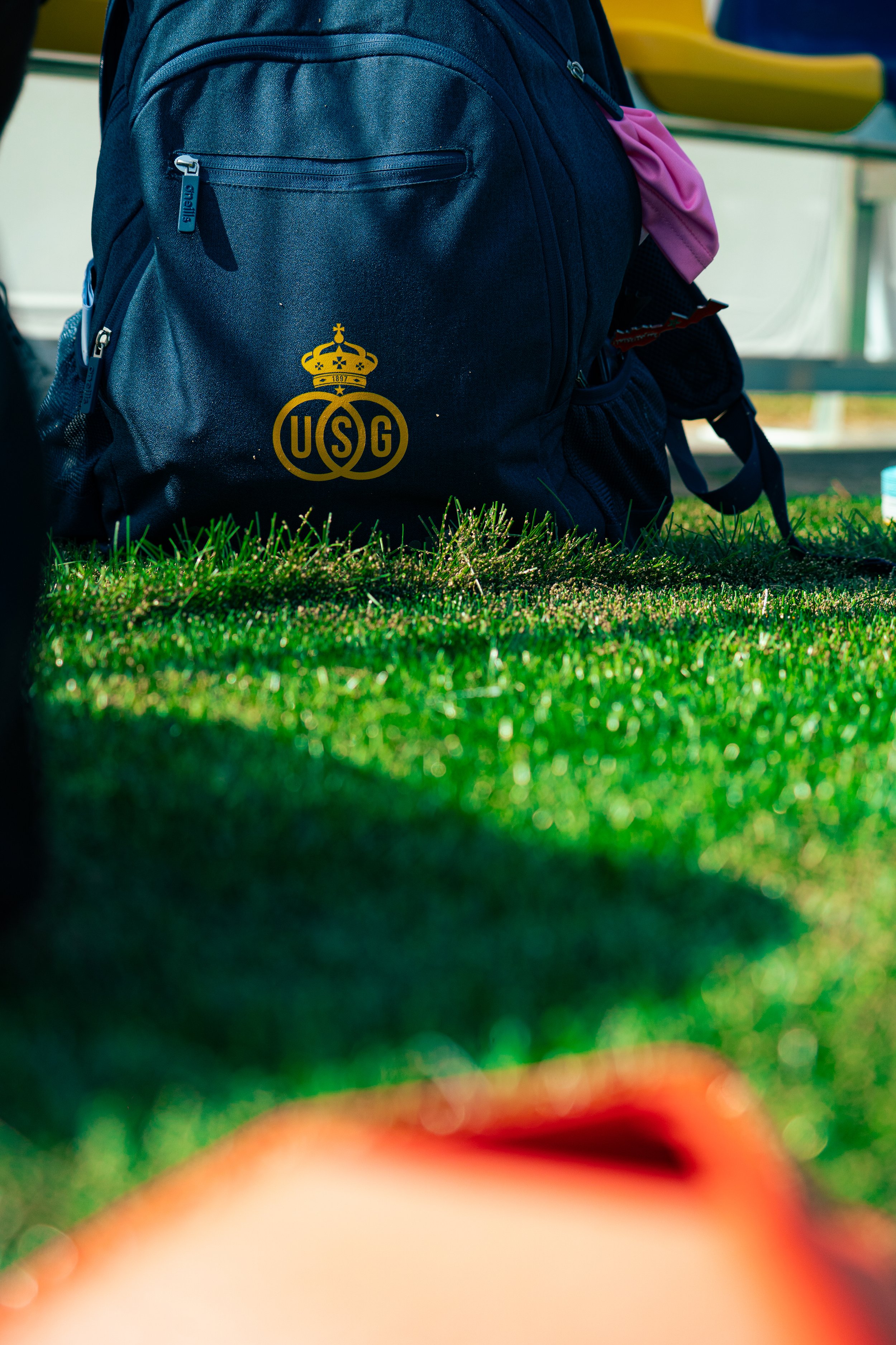 Close-up of a navy backpack with a yellow USG logo, resting on green grass, with part of a pink object and a bench in the background.