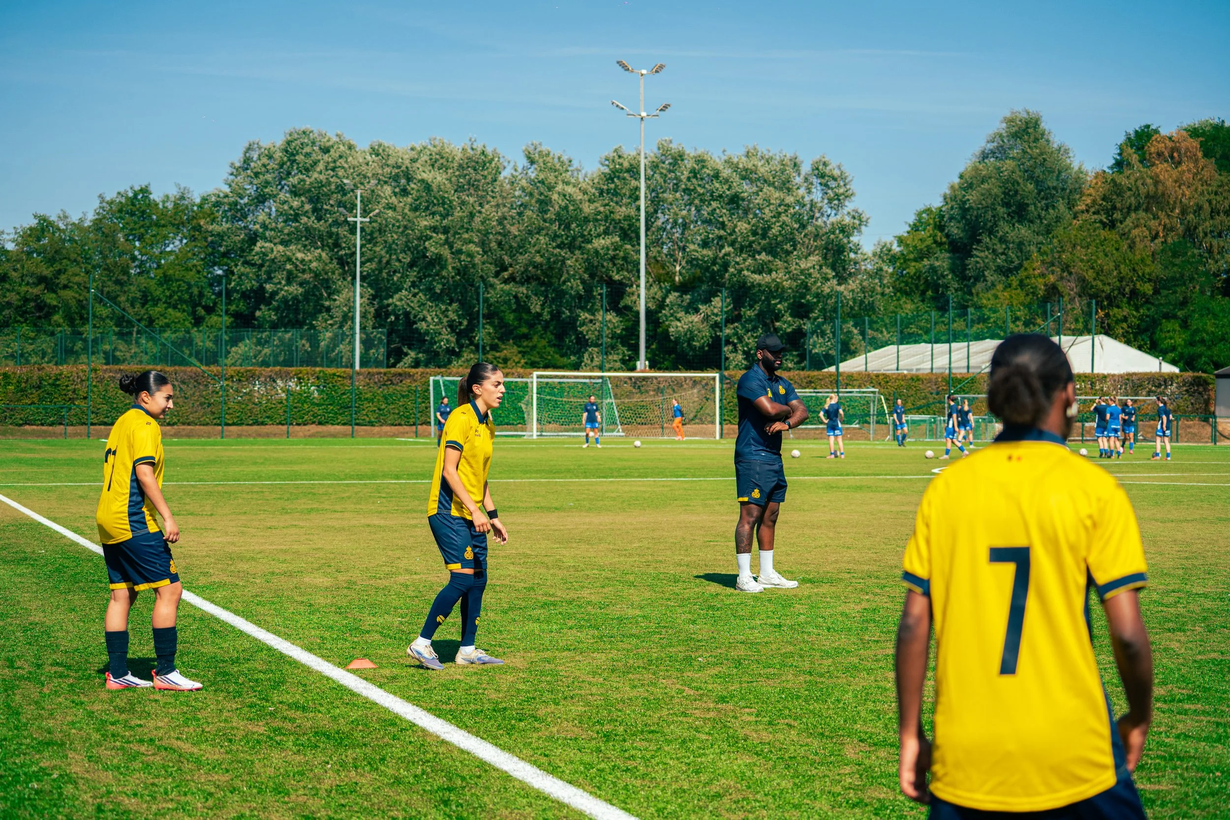 Soccer players in yellow and blue uniforms standing on a grass field during a practice or game, with coaches and other players visible in the background, trees, and clear blue sky.