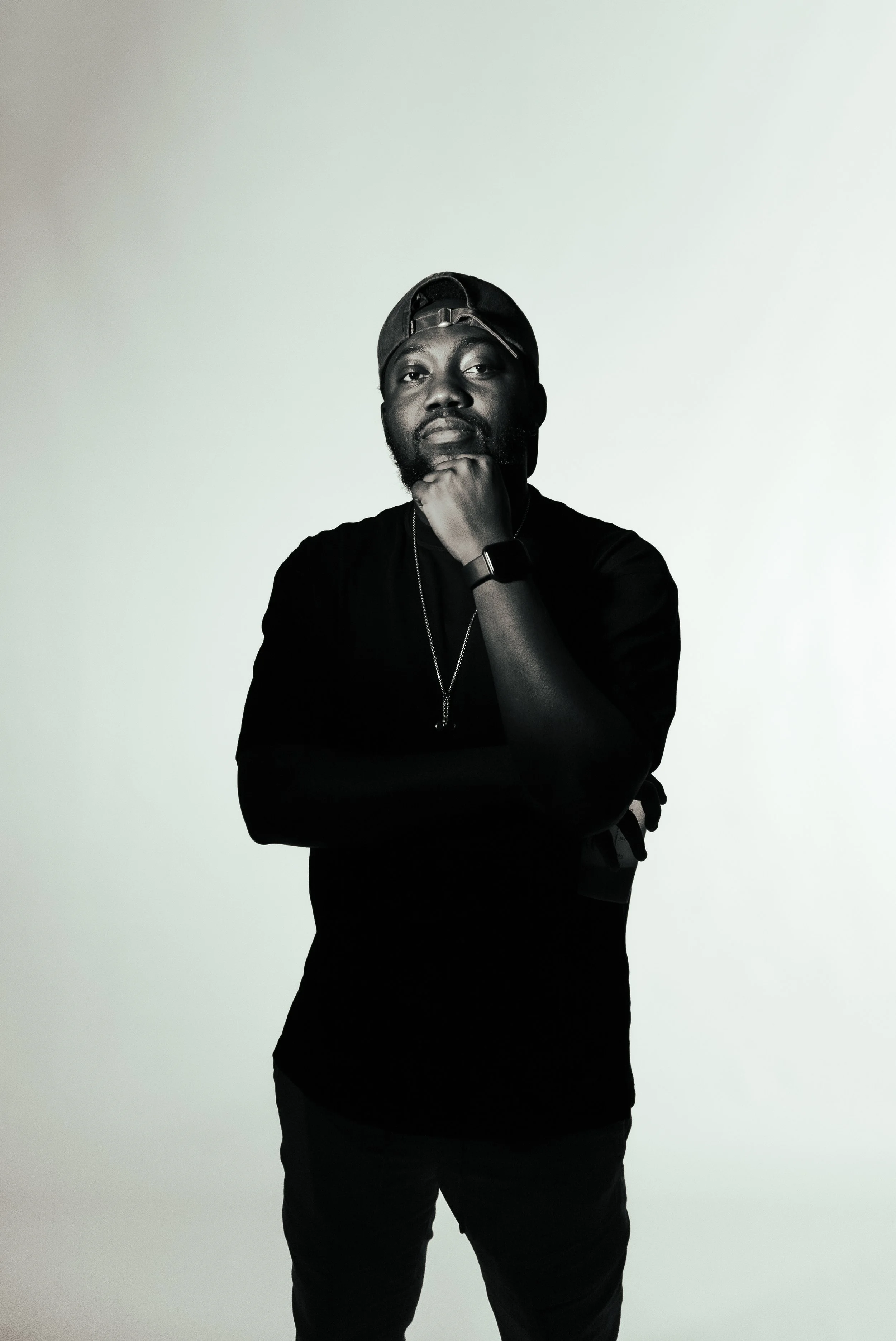 Portrait of a man wearing a backward black cap, black t-shirt, and a smartwatch, standing against a plain light-colored background. He poses with his right hand under his chin and looks serious.