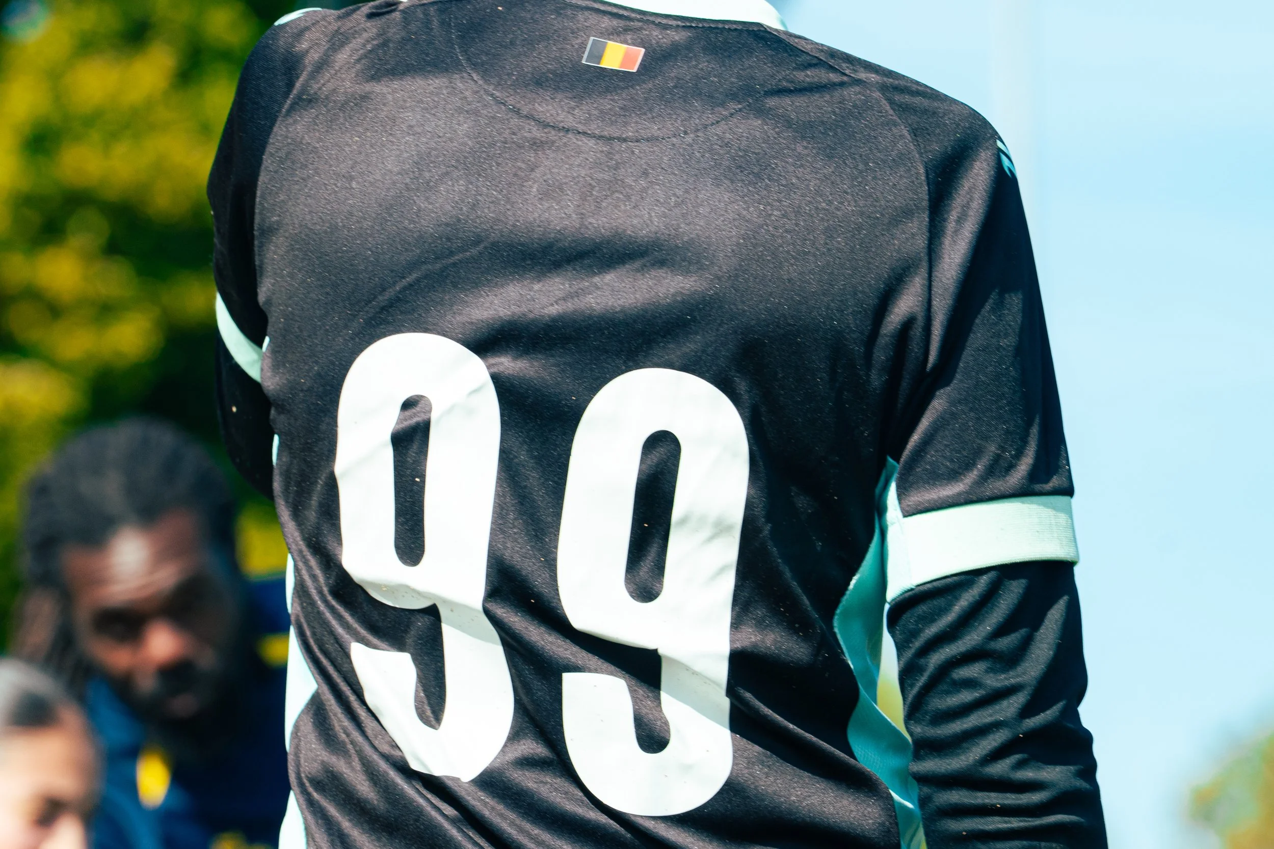 Close-up of a sports jersey with a Belgian flag pin attached, worn by a person during a game outdoors.