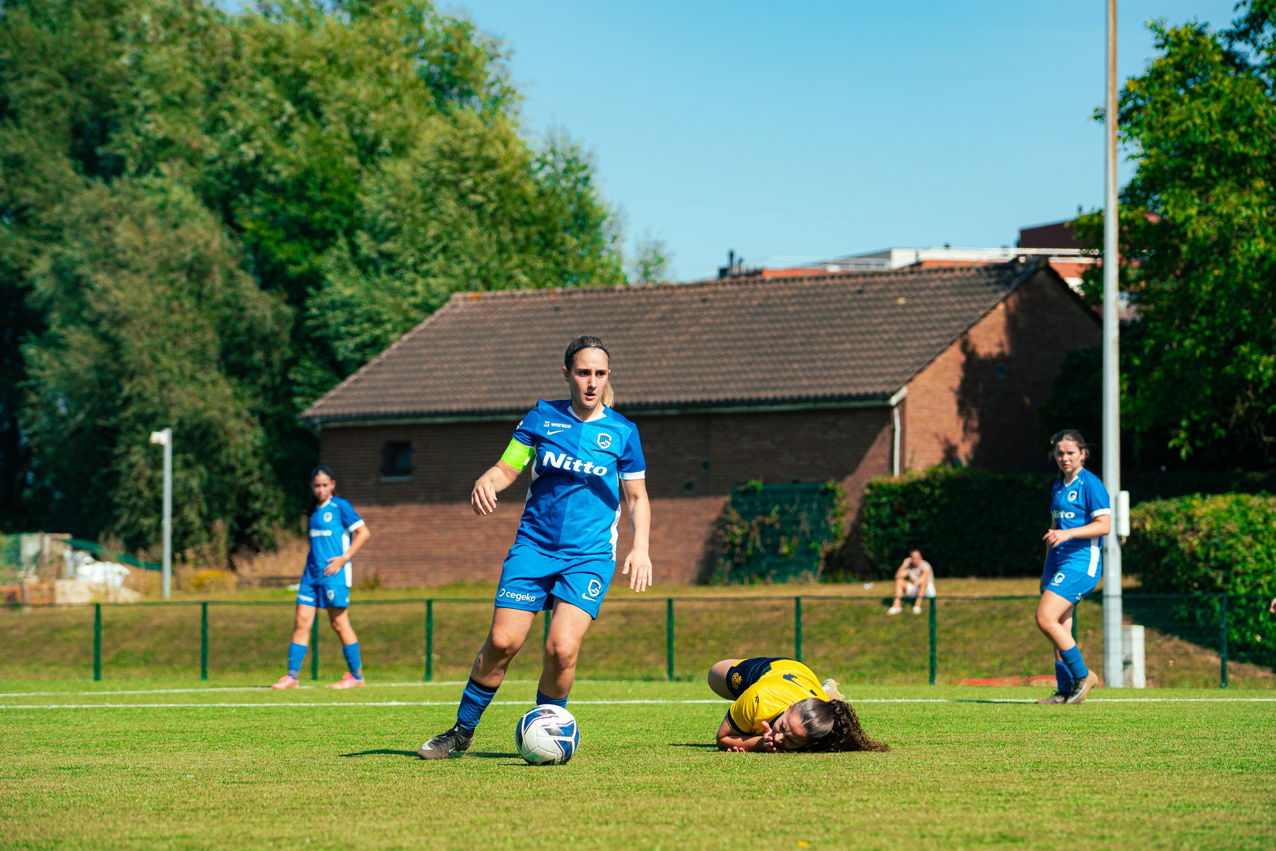 A women's soccer match with players in blue jerseys on a grassy field, one player standing over a fallen opponent in a yellow jersey, with a background of trees, houses, and clear blue sky.