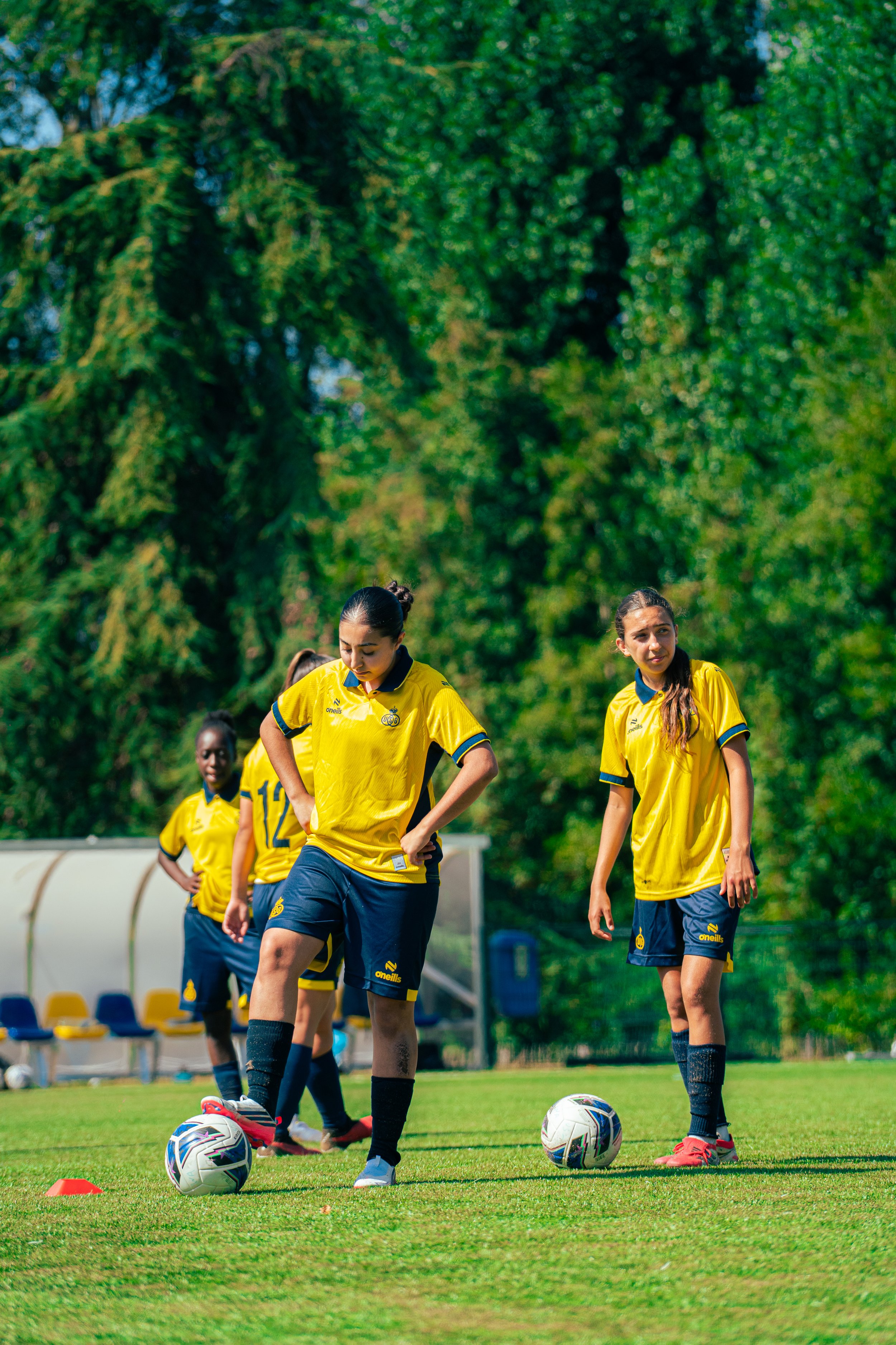 Women in yellow and navy soccer uniforms practicing on a grassy field, with soccer balls and a row of chairs in the background, surrounded by trees.