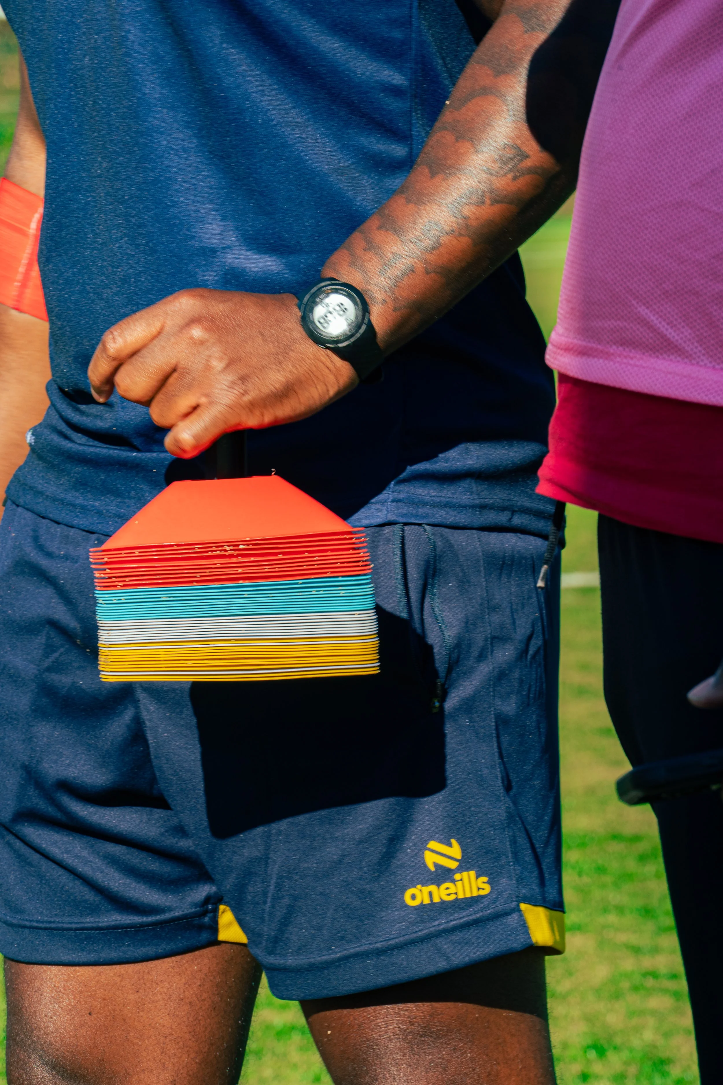 Close-up of two rugby players on a field, one wearing dark blue O'Neills shorts and the other wearing a pink jersey. The player in dark shorts is holding a stack of colorful and white cards.