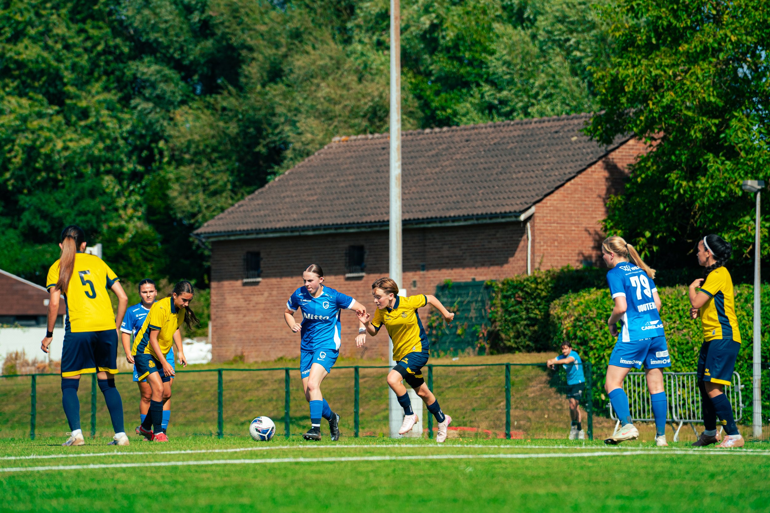 Women and girls playing soccer on a green field, wearing yellow and blue jerseys, with trees and houses in the background.