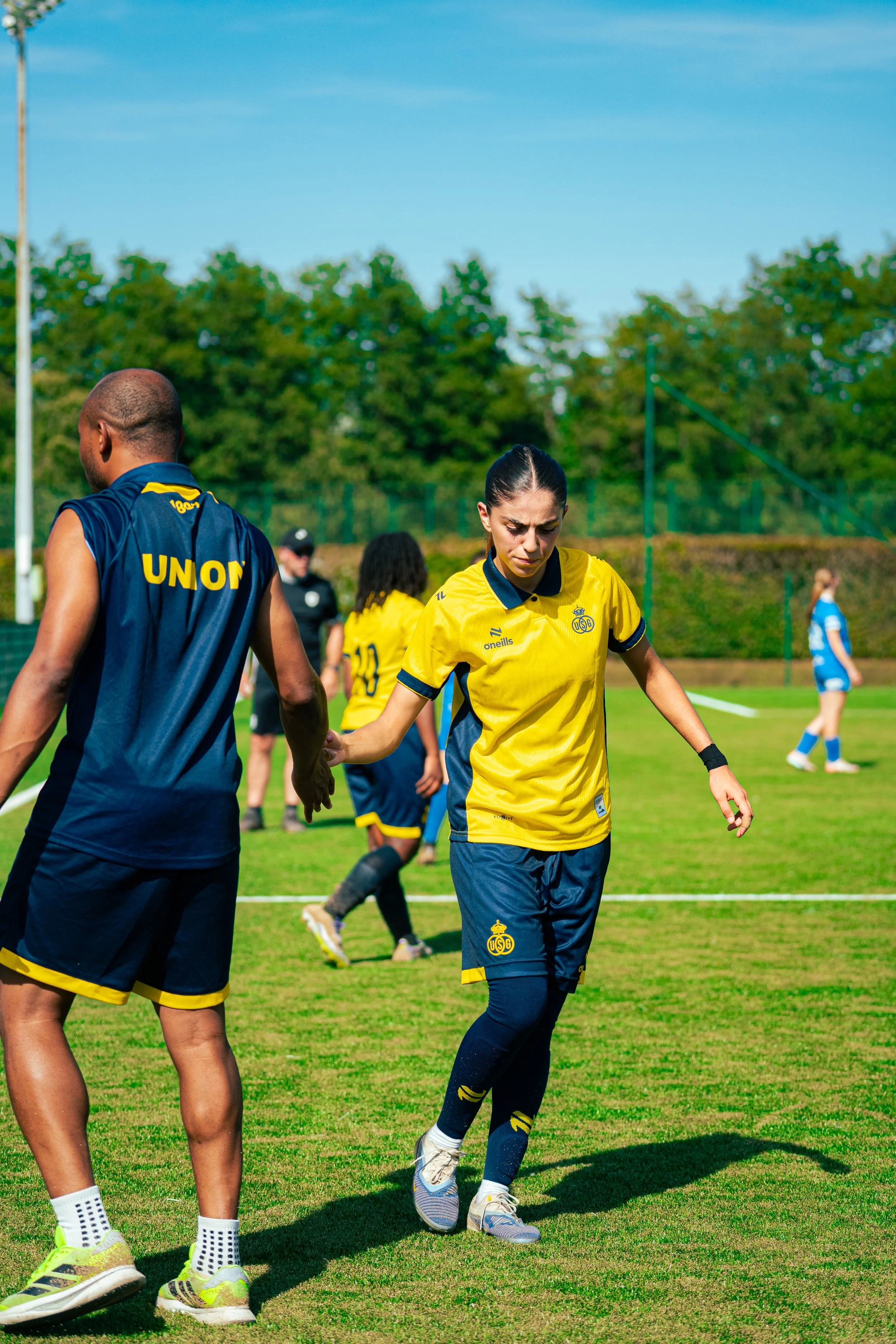 A group of soccer players on a green field during daytime, handshaking after a match, with blue skies and trees in the background.