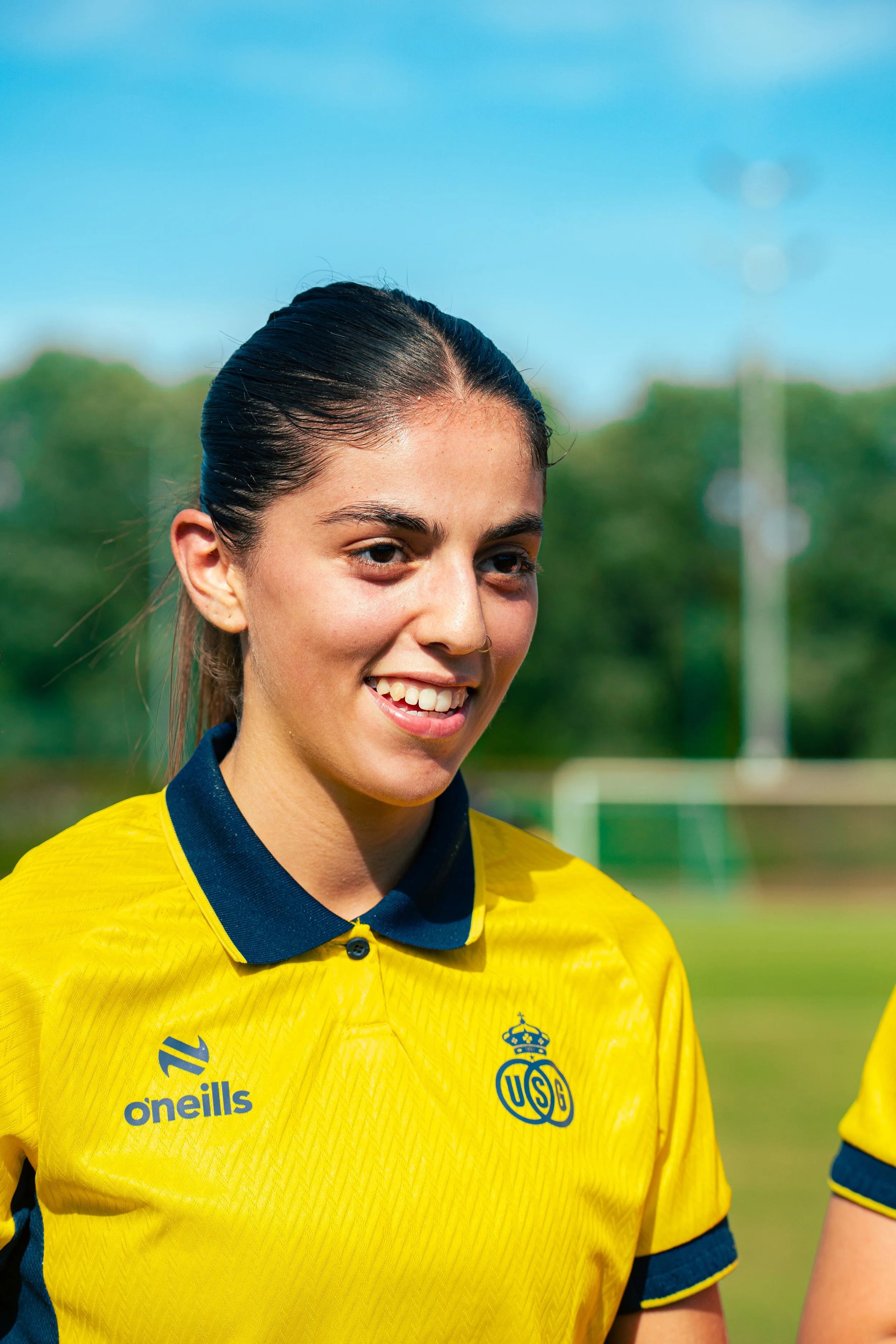 Close-up of a young woman wearing a yellow soccer jersey with a navy collar, smiling, standing outdoors on a sunny day with a blue sky and green trees in the background.