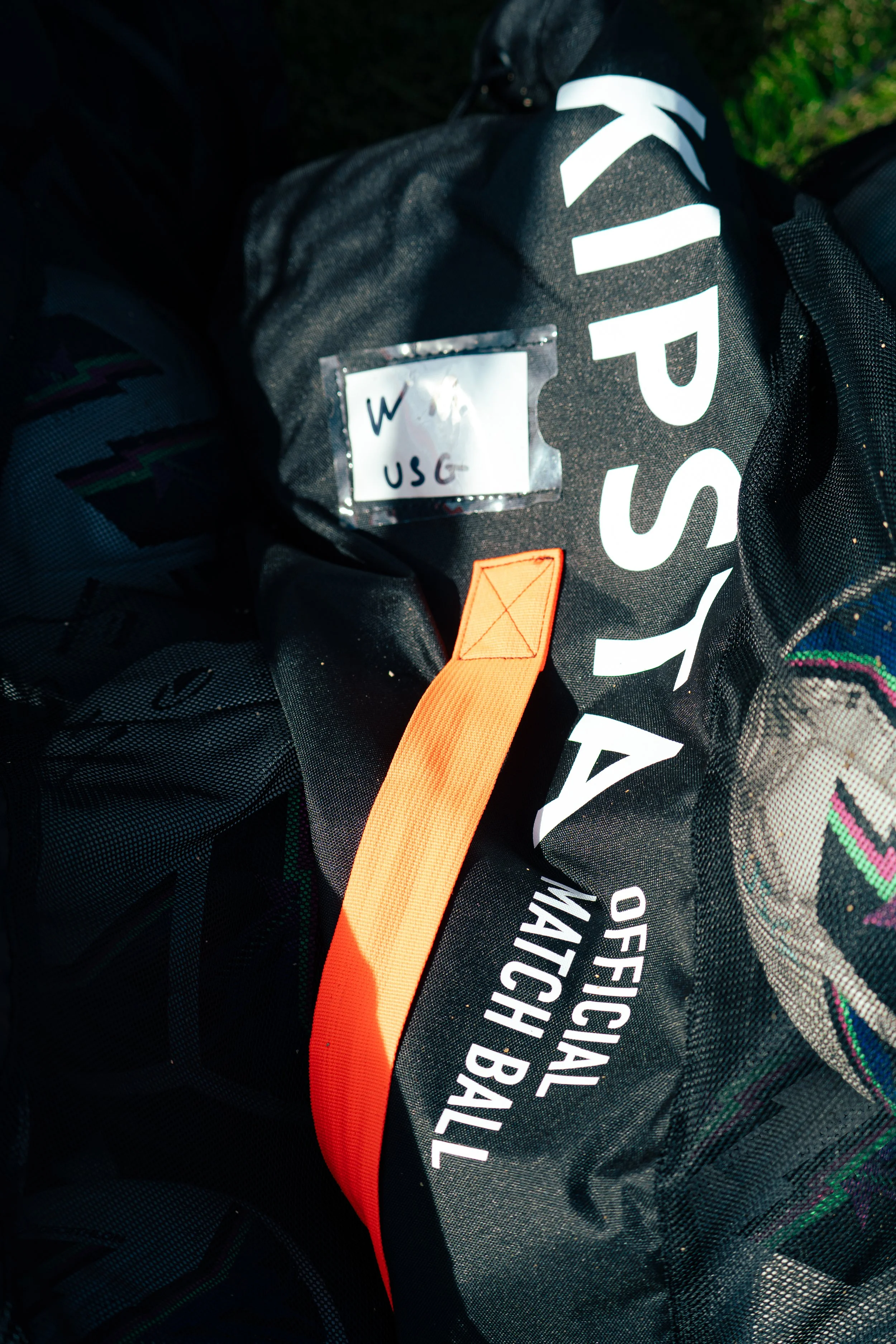 Close-up of a black backpack with white text and orange and red straps, placed on grass at night.